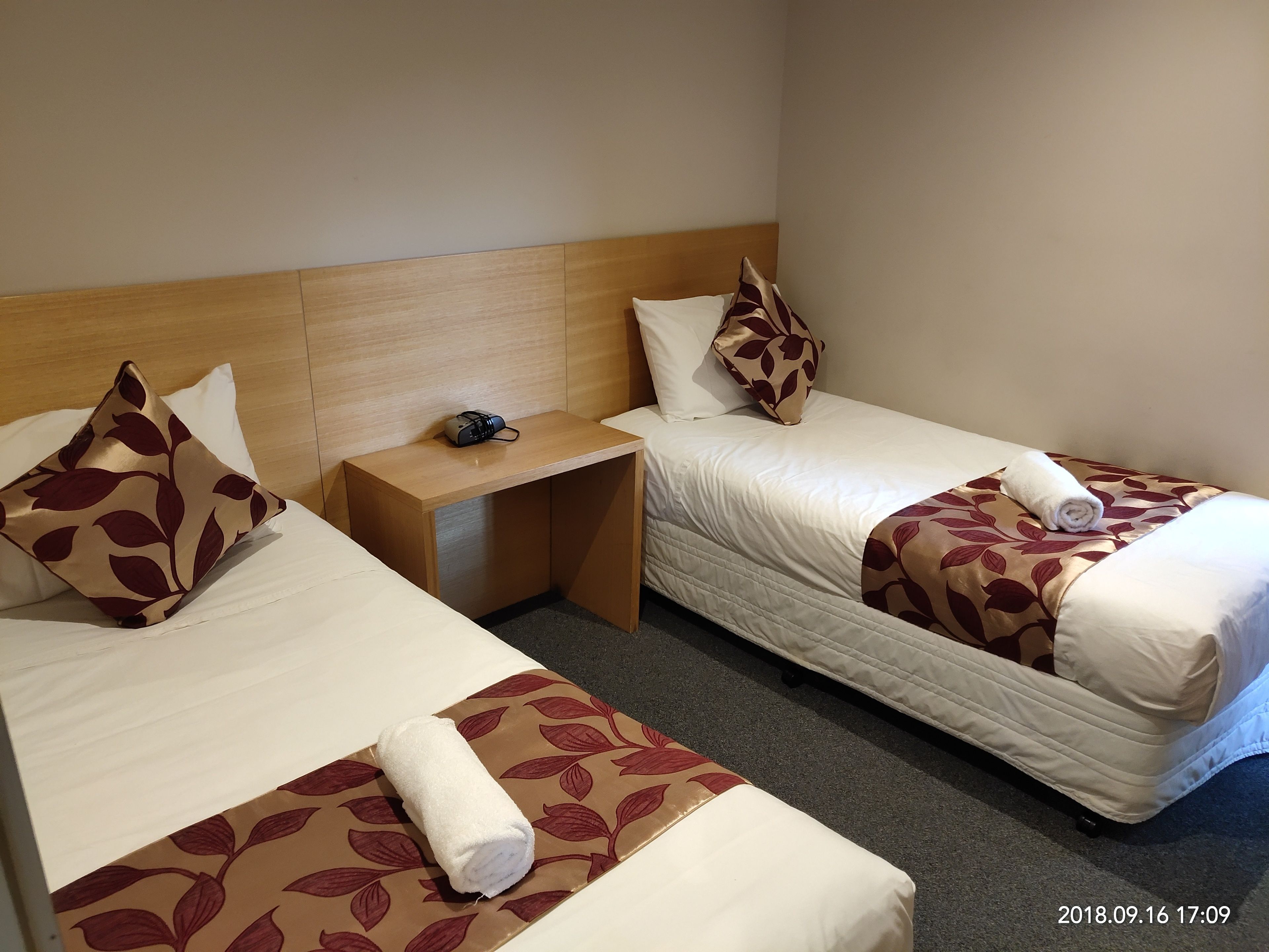 A hotel room with two single beds, each with a pillow and a rolled towel. There is a small wooden nightstand between the beds with a lamp and a small electronic device.