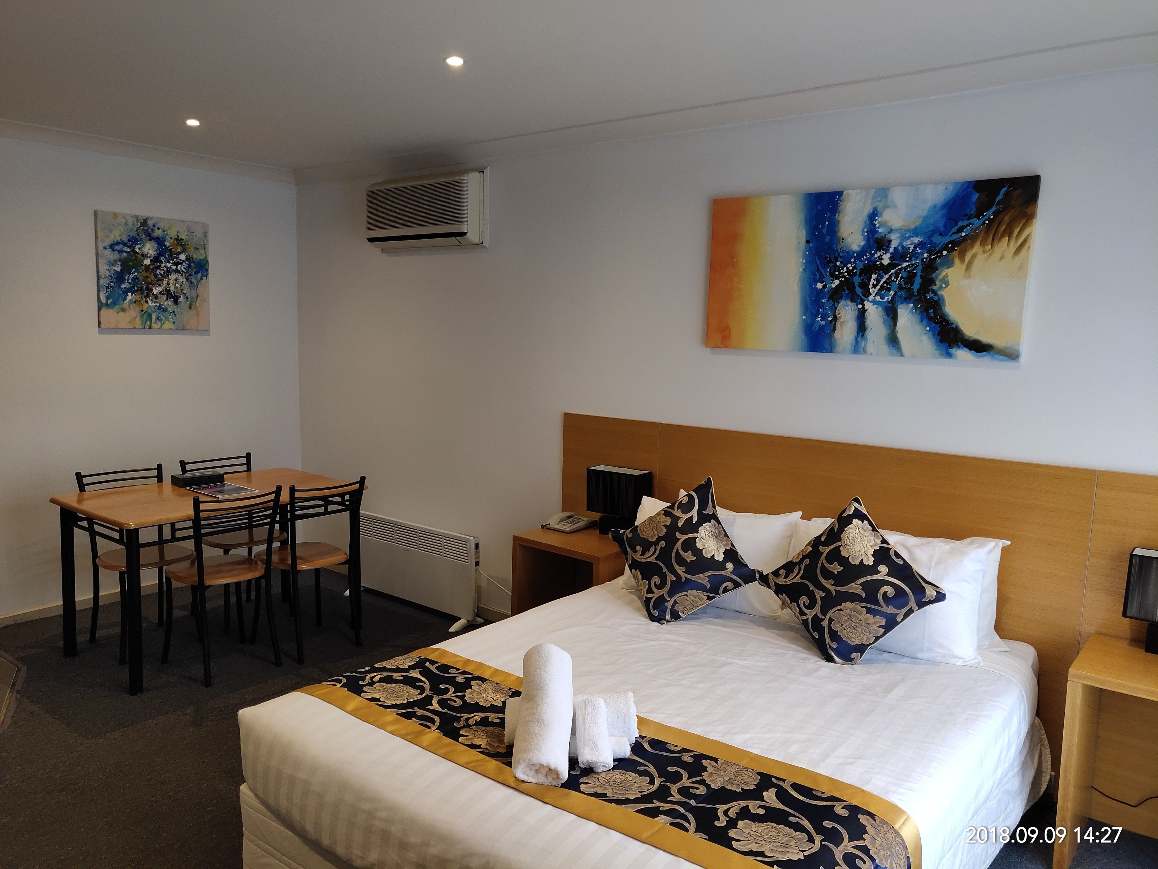 A hotel room with a bed, a desk with chairs, a painting on the wall, and an air conditioning unit.