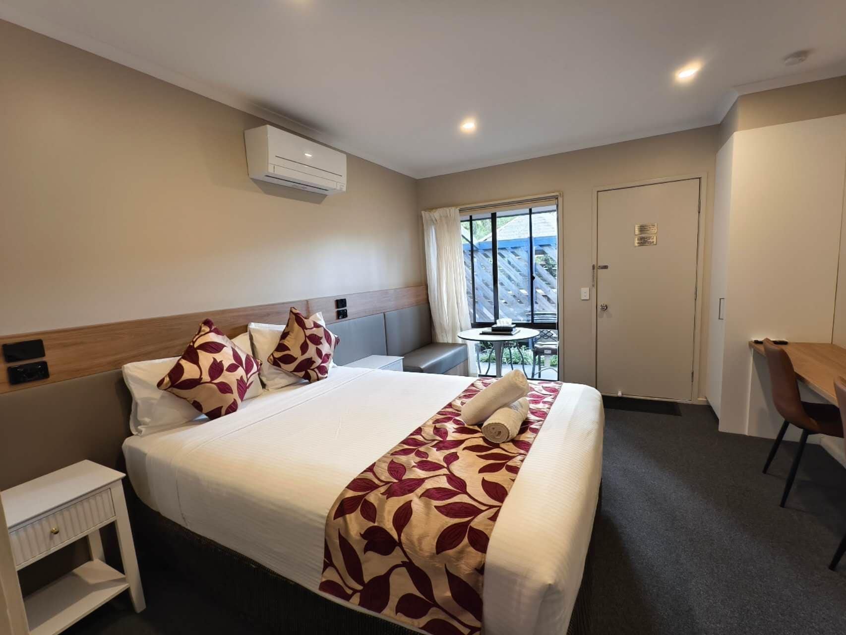 Een hotelkamer met een bed, nachtkastje, stoel, en een klein tafeltje met een stoel voor het raam.