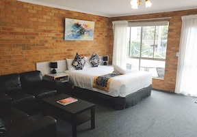 Habitación de hotel con pared de ladrillo, una cama grande con almohadillas estampadas, un sofá de cuero negro, una mesita pequeña y una ventana con cortinas.
