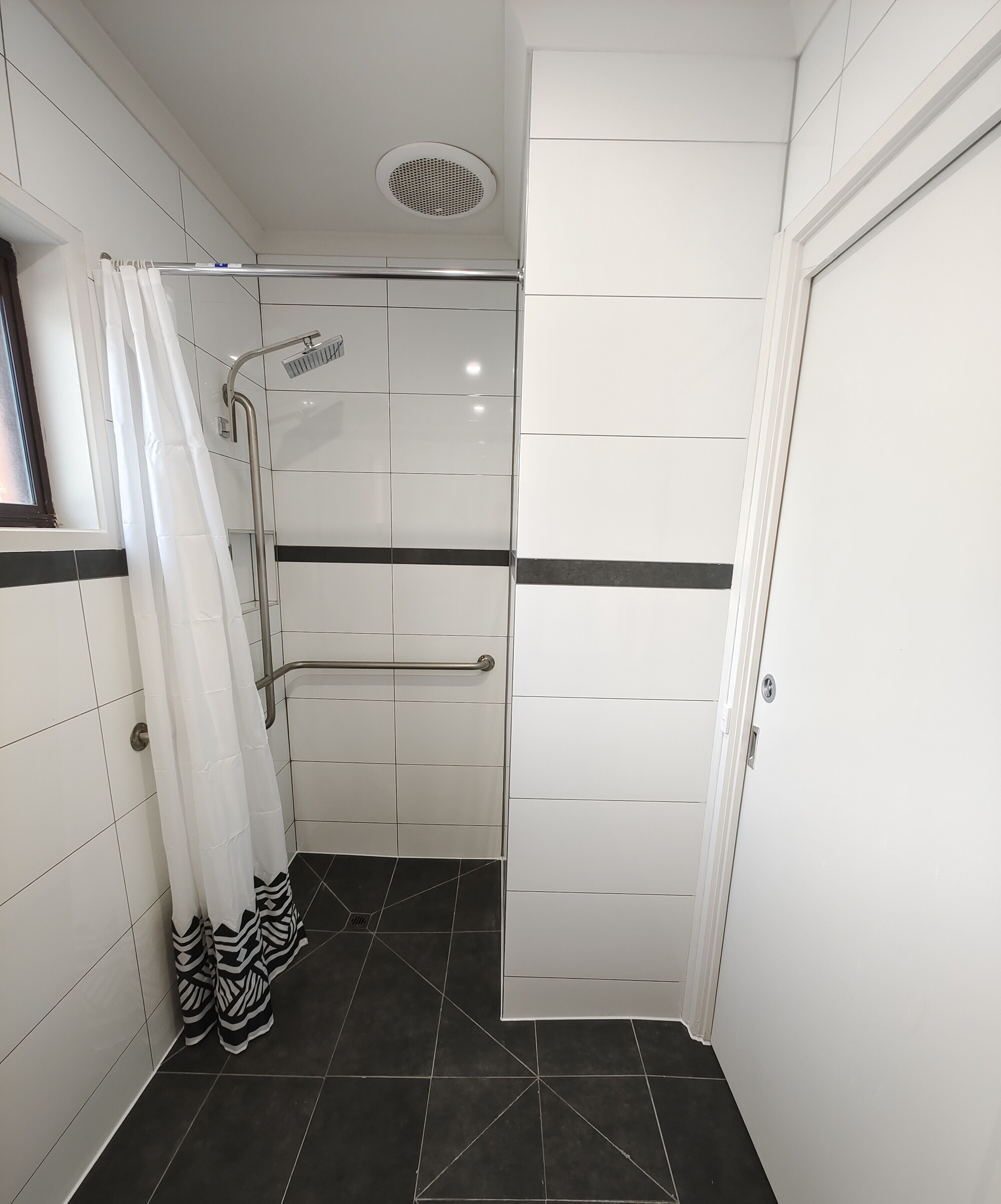A bathroom with white tiled walls, a shower with a handheld showerhead, a black and white patterned shower curtain, and a door with a handle.