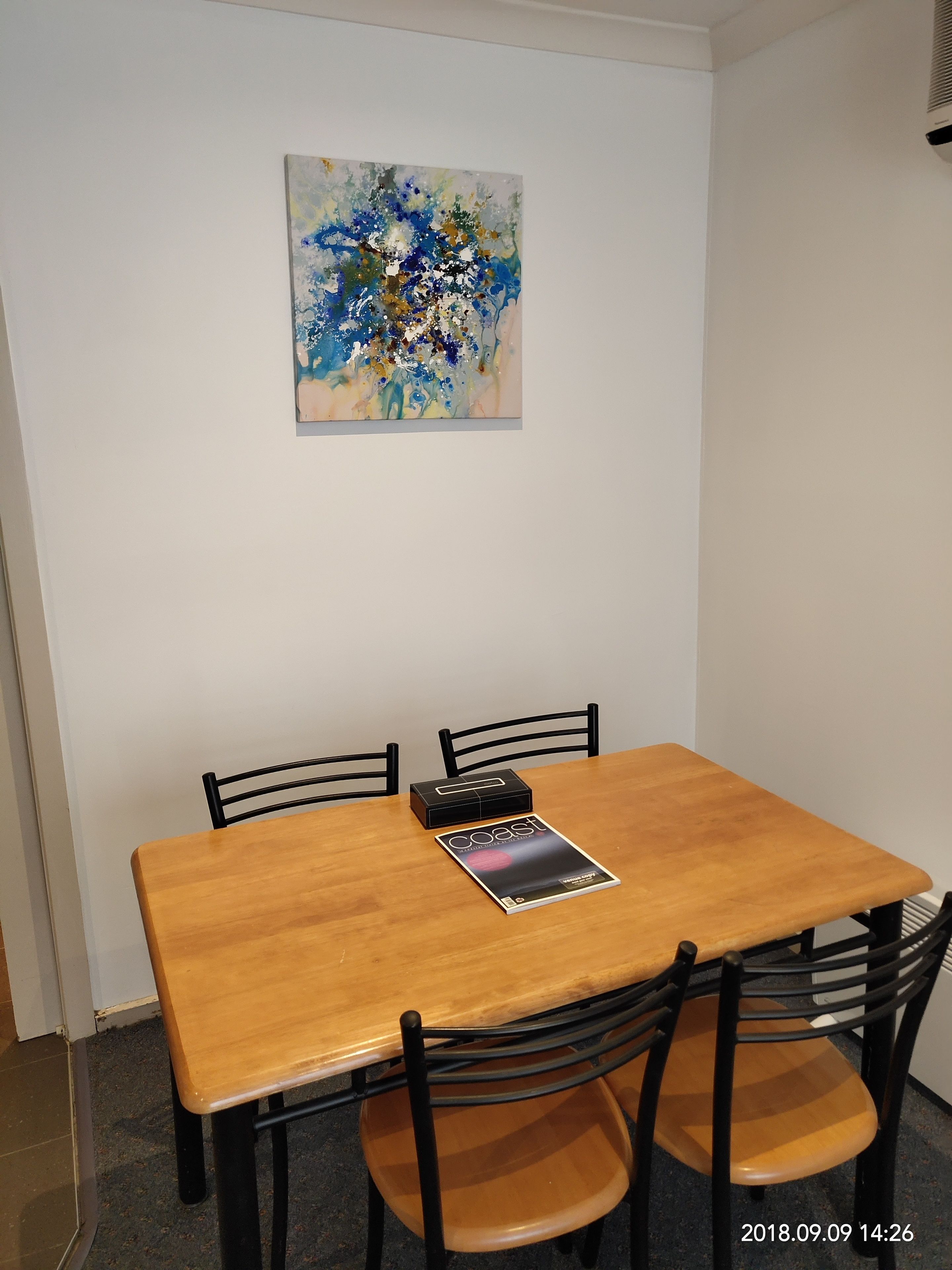 A dining area with a wooden table, four chairs, and a painting on the wall.