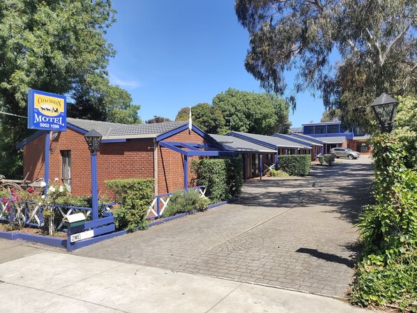 A motel with a brick building and blue awnings, a signboard, and a paved driveway. - The Coachman Motel & Units (Cowes)