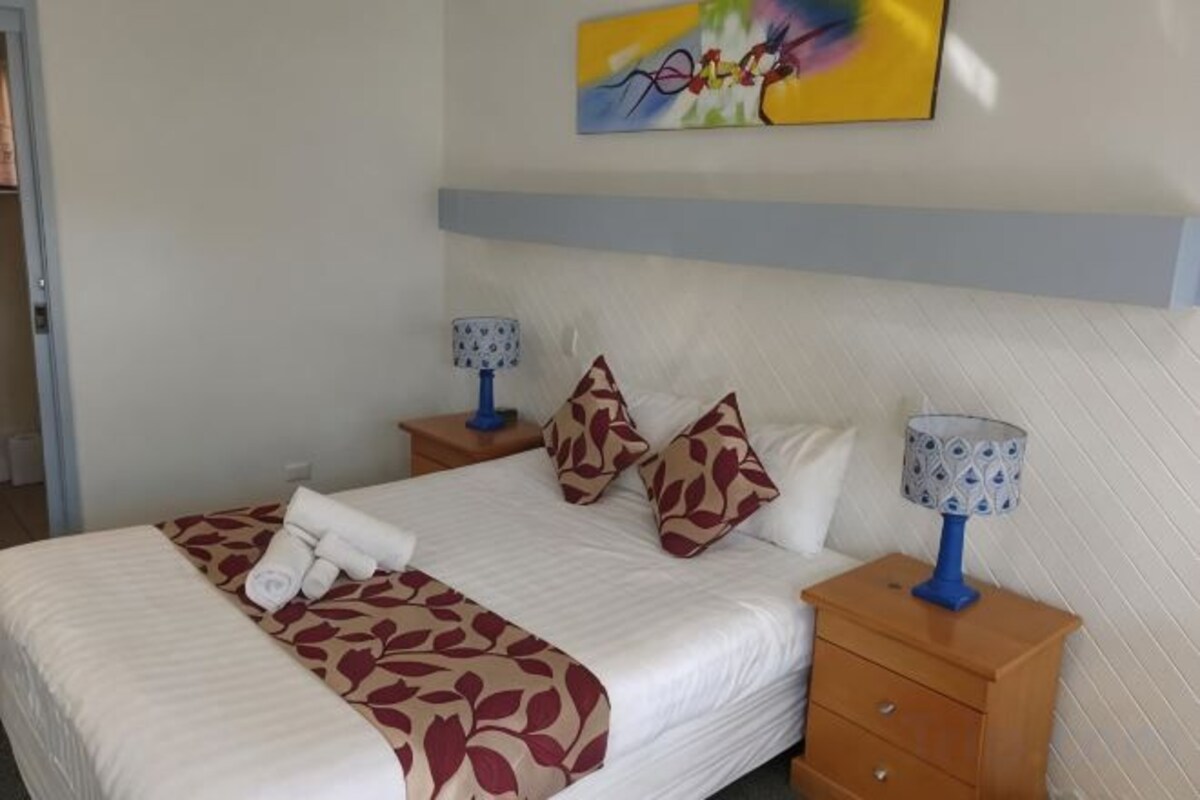 A neatly made bed with a patterned bedspread, two pillows, and a rolled towel. There are two bedside tables with lamps, and a wall-mounted artwork above the bed.