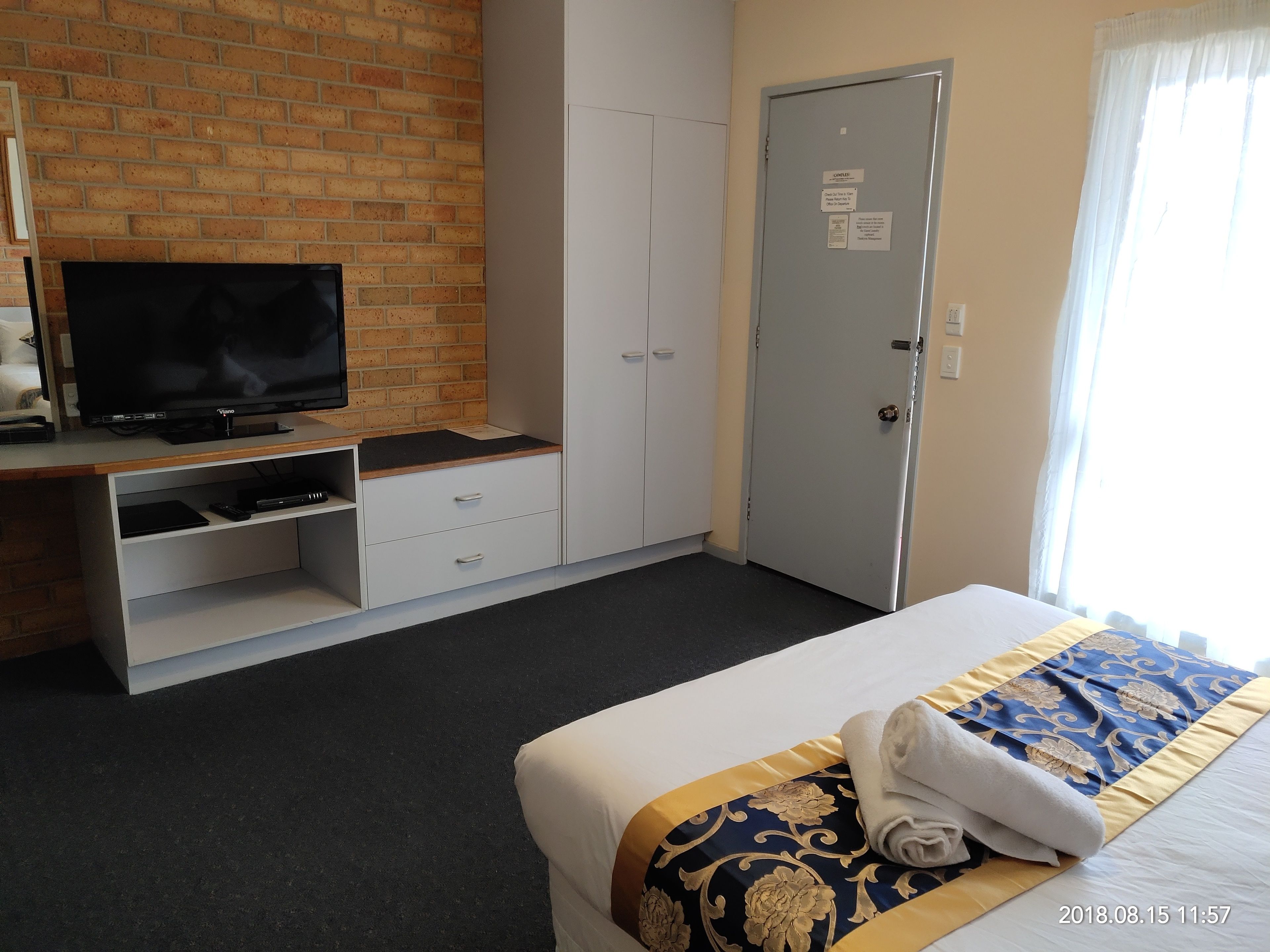 Habitación de hotel con una televisión sobre un soporte de madera, un armario, una puerta con un cartel y una cama con una manta estampada.