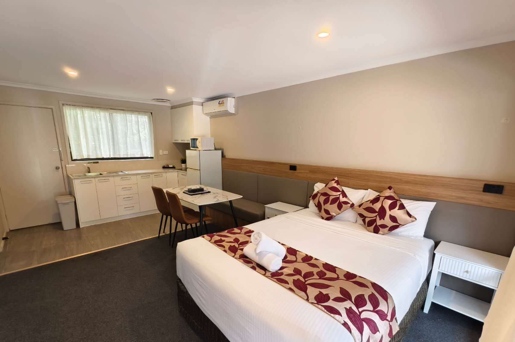 Ein Hotelzimmer mit Bett, Nachttisch, kleinem Schreibtisch mit Stuhl, Fenster mit Vorhängen und einer Küchenzeile.