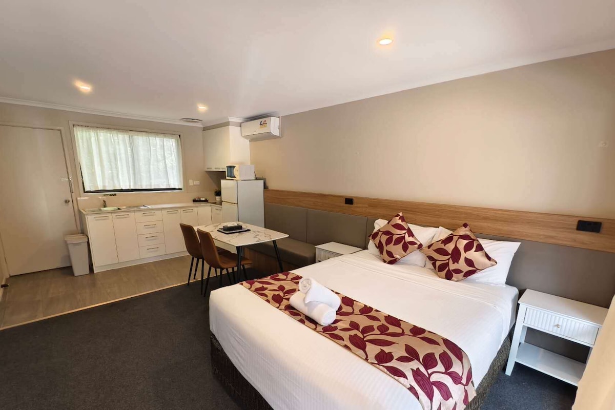 Habitación de hotel con cama, mesita de noche, un pequeño escritorio con silla, ventana con cortinas y una zona de kitchenette.