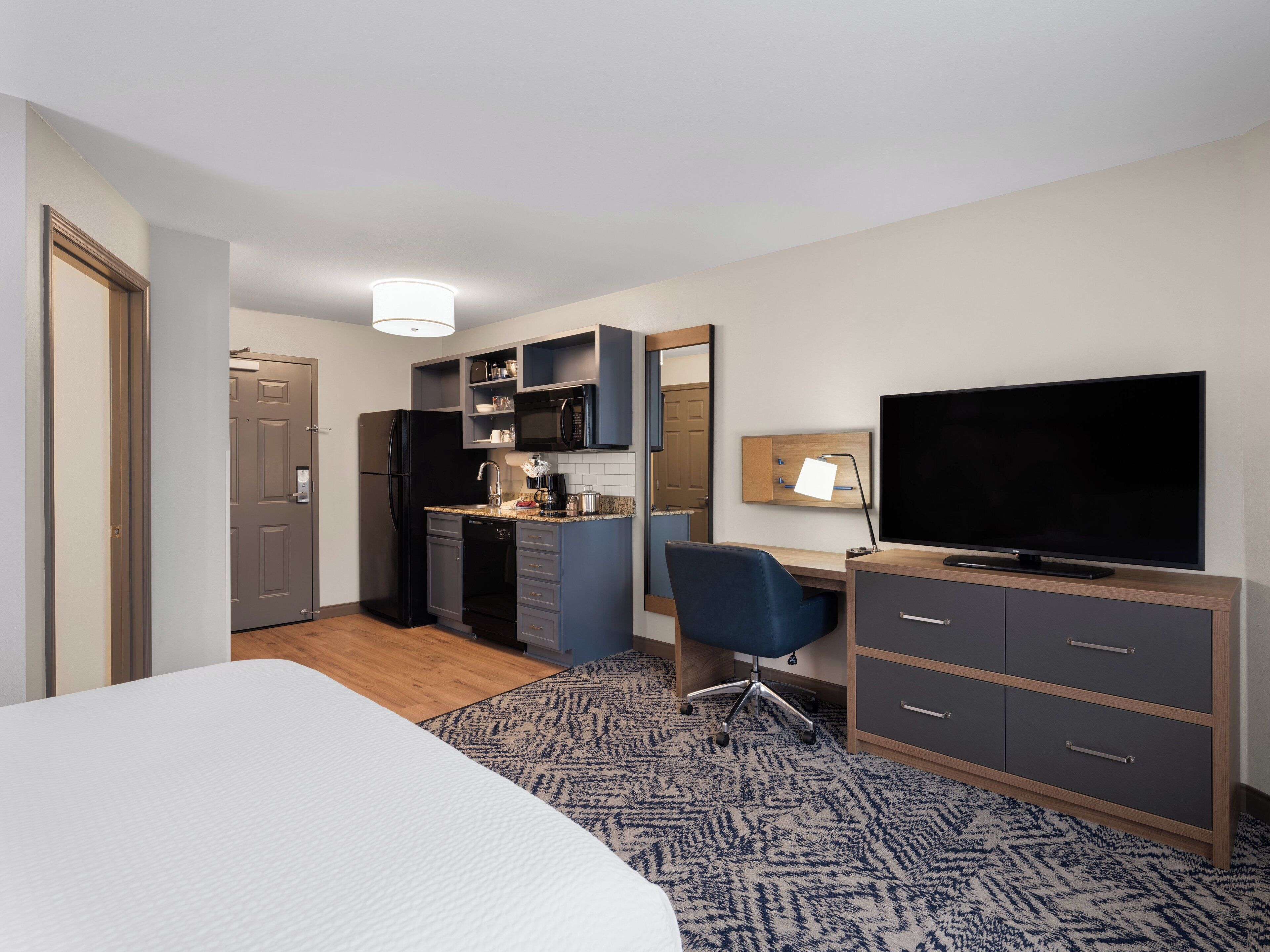 A hotel room with a bed, a desk with a computer, a TV, a kitchen area with a fridge, microwave, and sink.