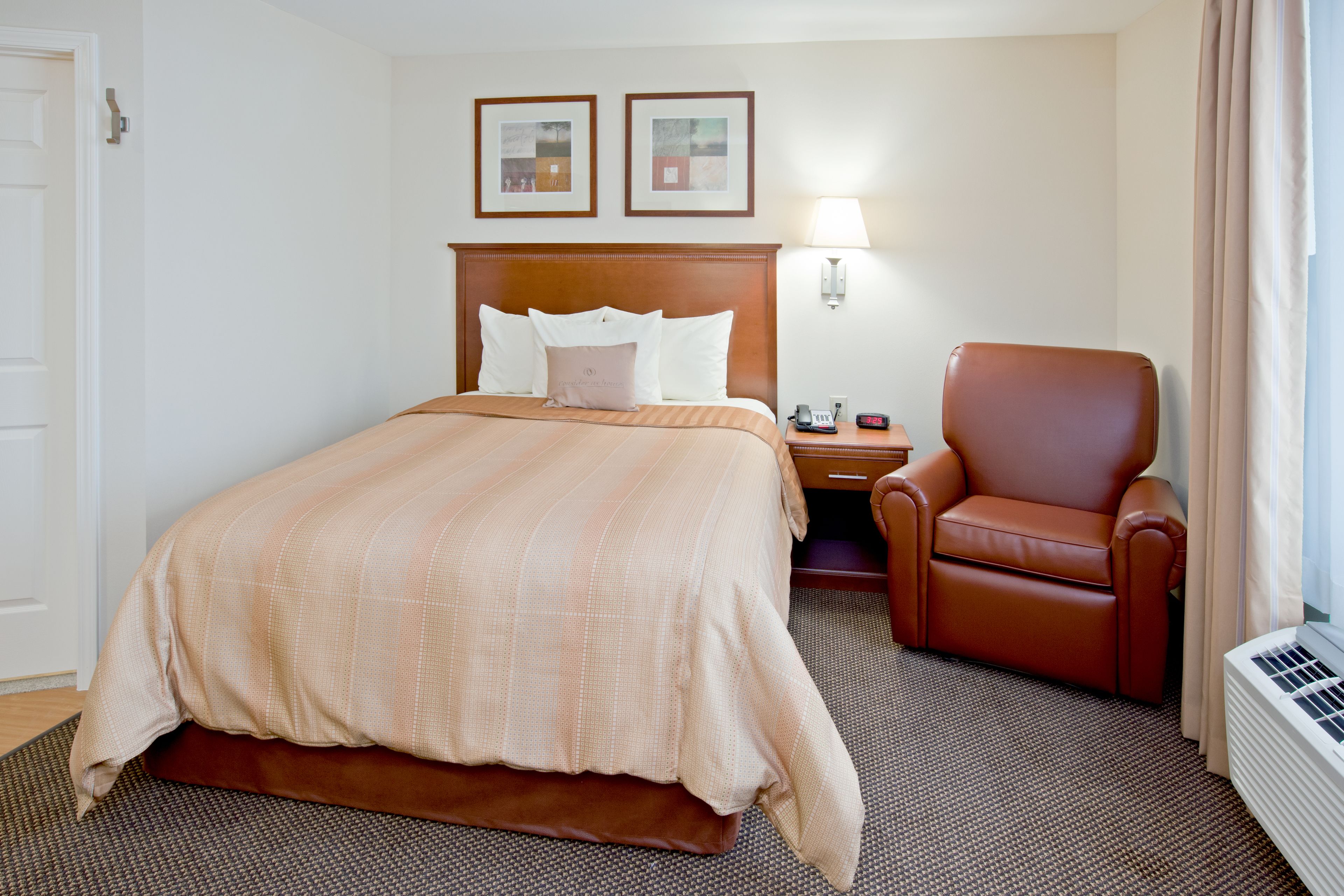 A hotel room with a large bed, a brown leather chair, a nightstand, a lamp, and a wall-mounted heater.