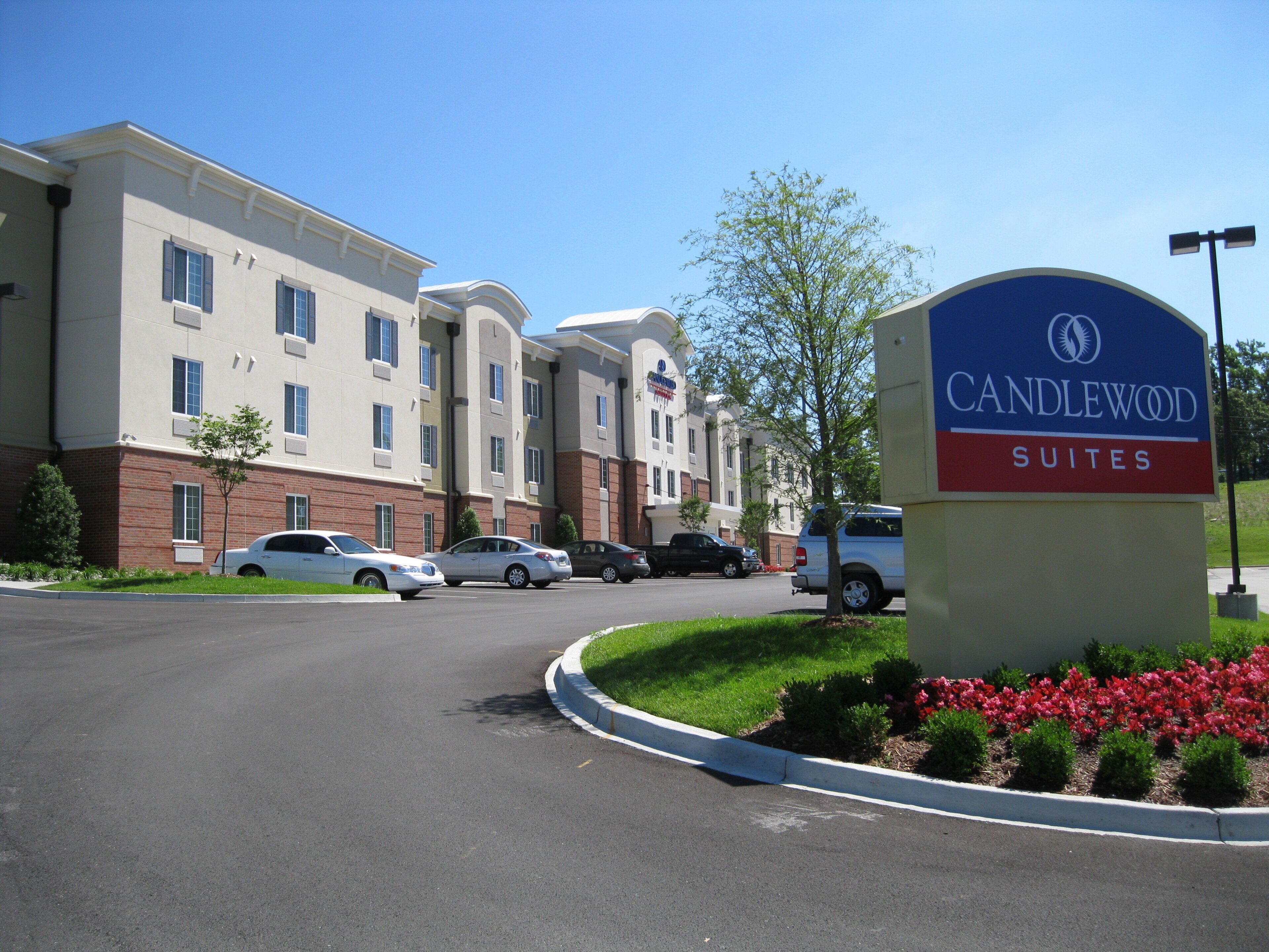A hotel entrance with a sign for Candlewood Suites, a parking lot with several cars, and a landscaped area with shrubs and flowers.
