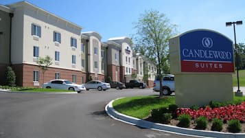 A hotel entrance with a sign for Candlewood Suites, a parking lot with several cars, and a landscaped area with shrubs and flowers.