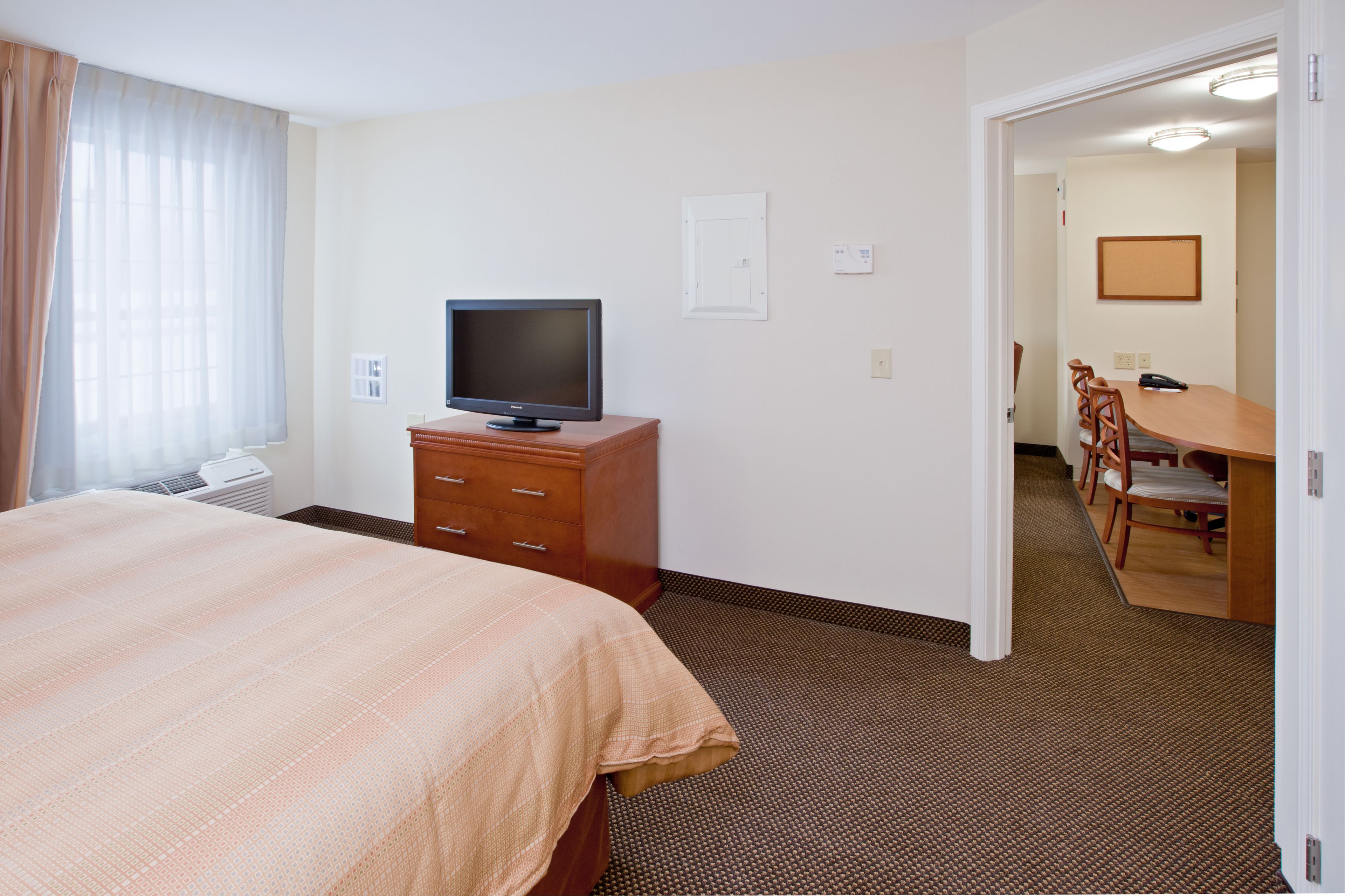 Habitación de hotel con cama, televisor sobre un soporte de madera, escritorio con sillas y zona de comedor.