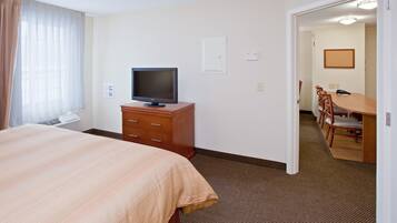 Habitación de hotel con cama, televisor sobre un soporte de madera, escritorio con sillas y zona de comedor.