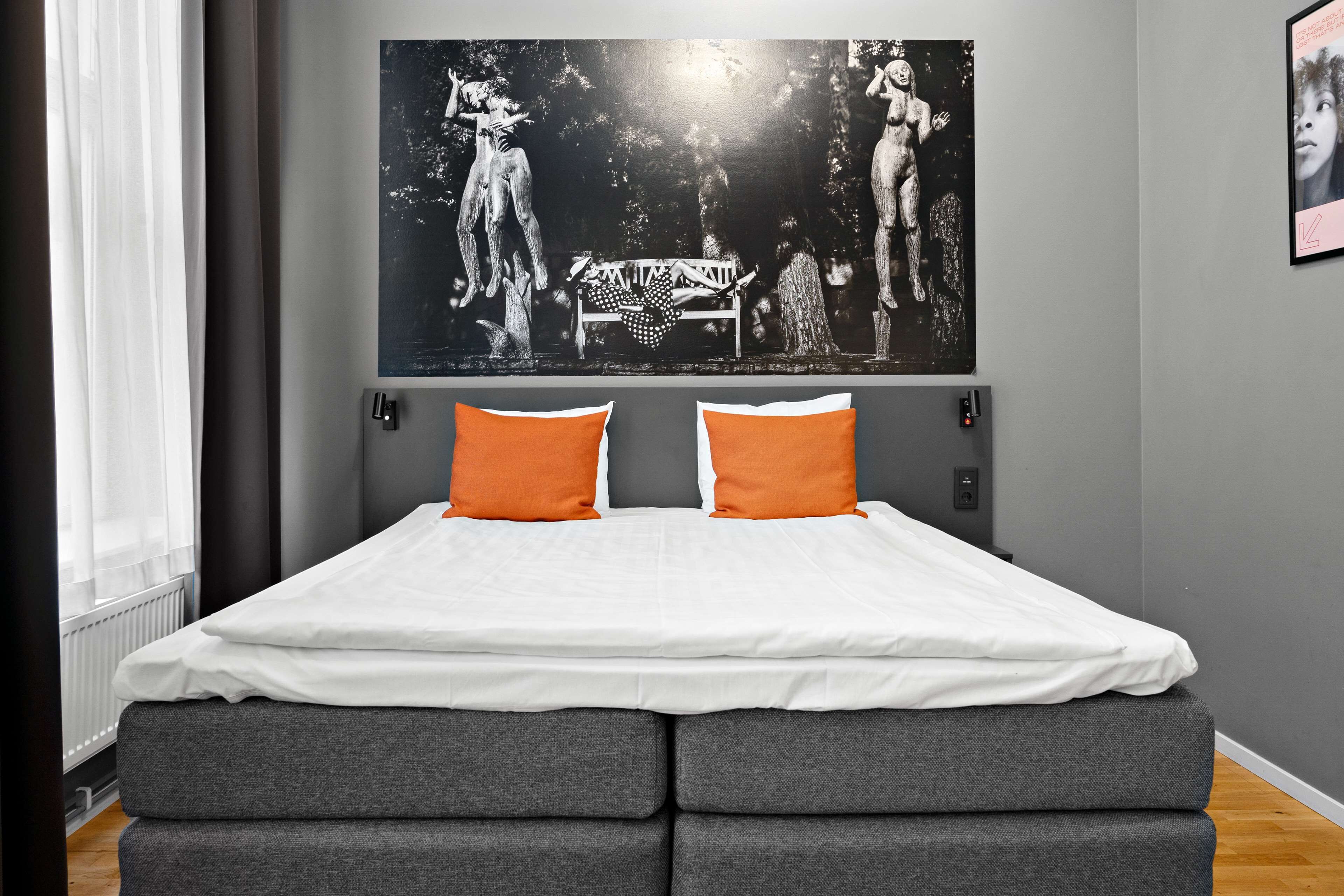 Un dormitorio con una cama grande, cabecera tapizada en gris, ropa de cama blanca y dos almohadas naranjas.