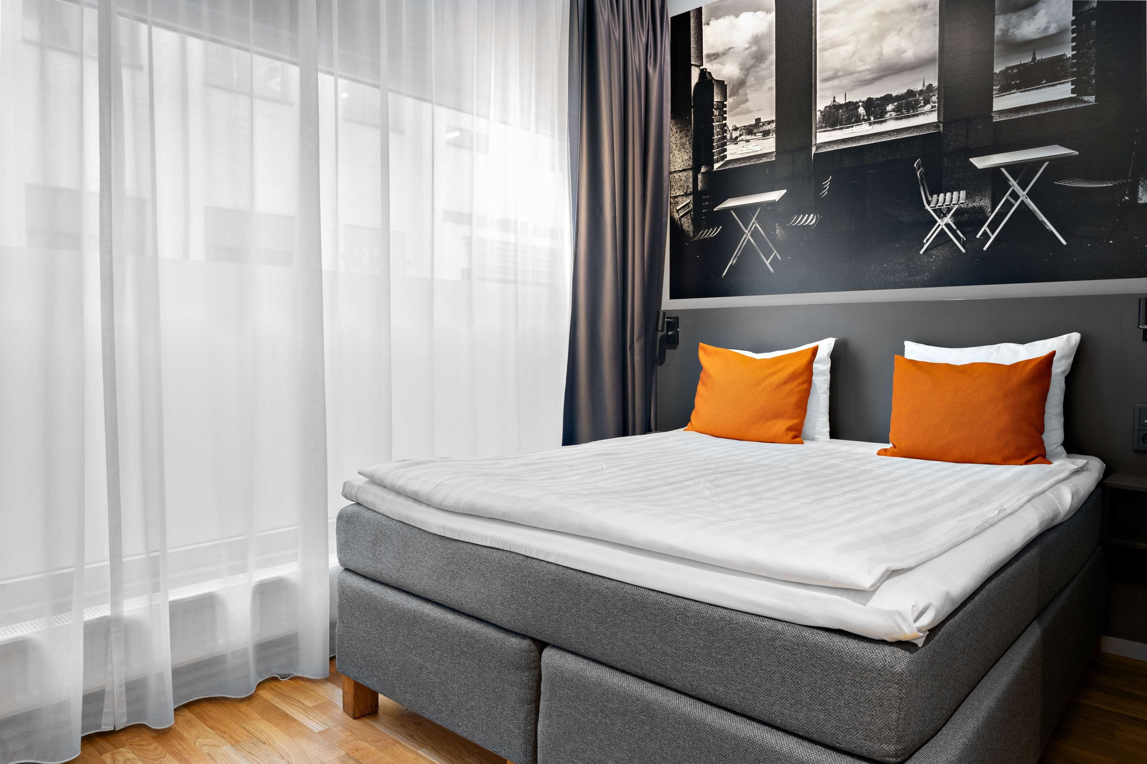 Quarto de hotel moderno com cama cinzenta, roupa de cama branca e almofadas laranja.