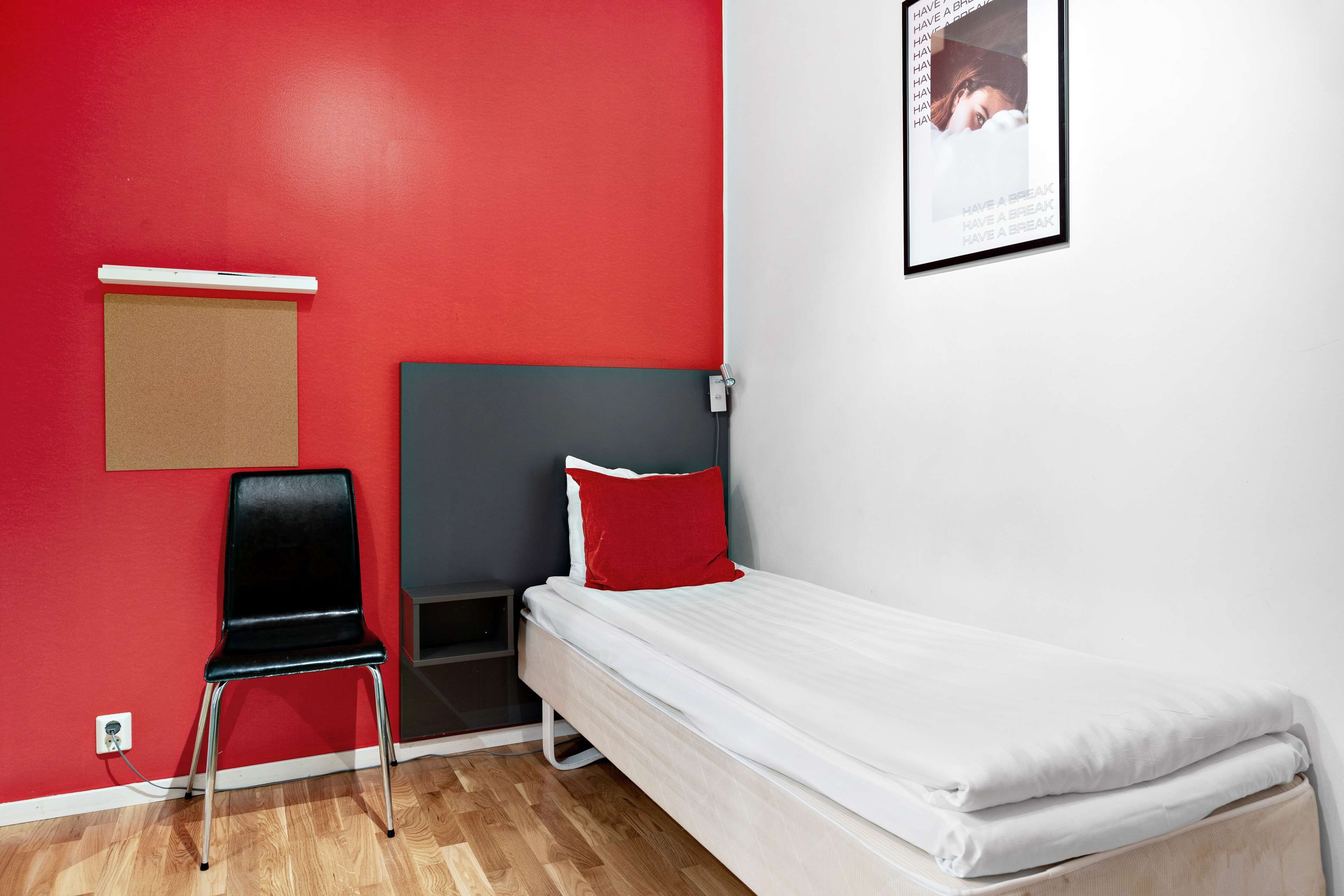 A small, single-bed room with a red wall, a black chair, and a white bed.