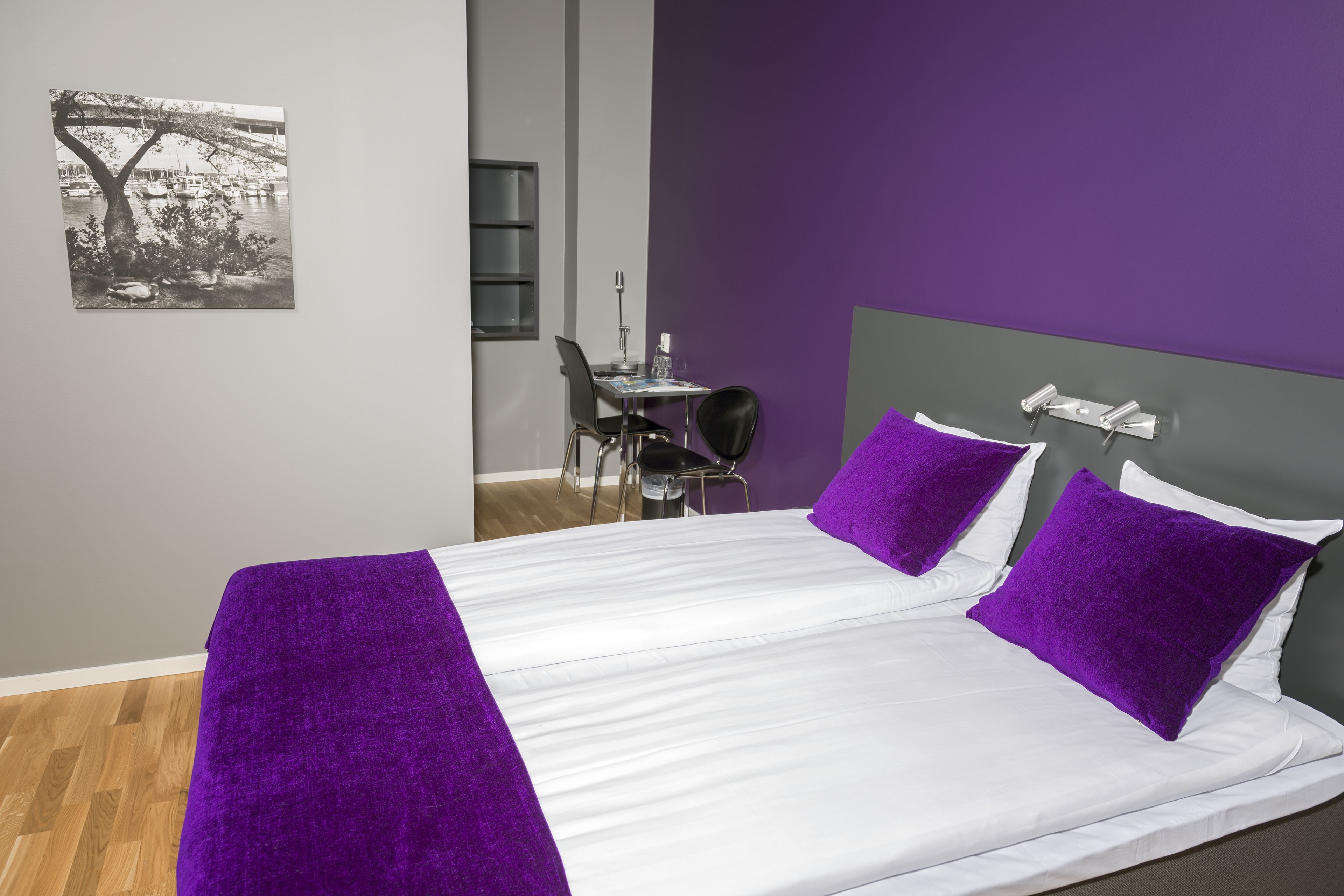 Un lit avec du linge de lit blanc et des touches de couleur violette, un petit bureau avec une chaise et une étagère fixée au mur.
