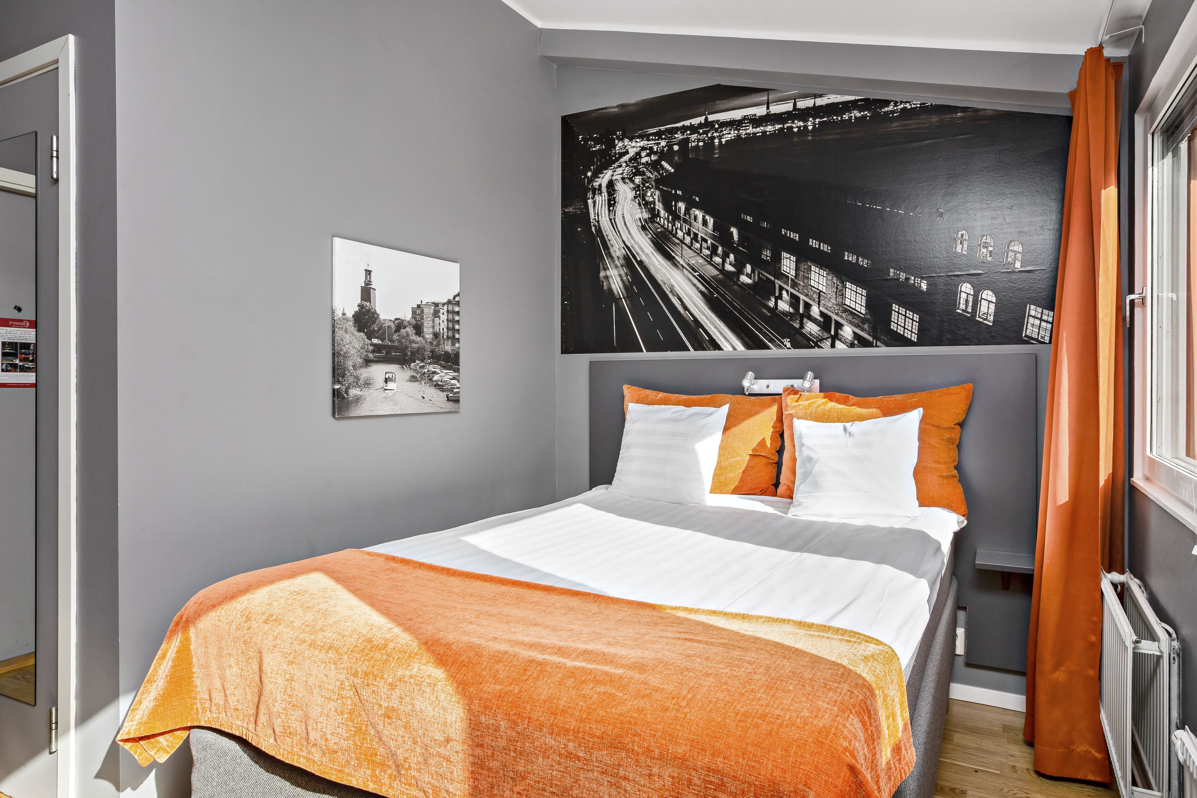 Ein modernes Hotelzimmer mit einem großen Bett, einem Stadtbild-Wandgemälde und Blick auf die Stadt durch das Fenster.