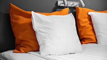 Una cama con almohadas blancas y anaranjadas, una pared gris y una lámpara moderna.