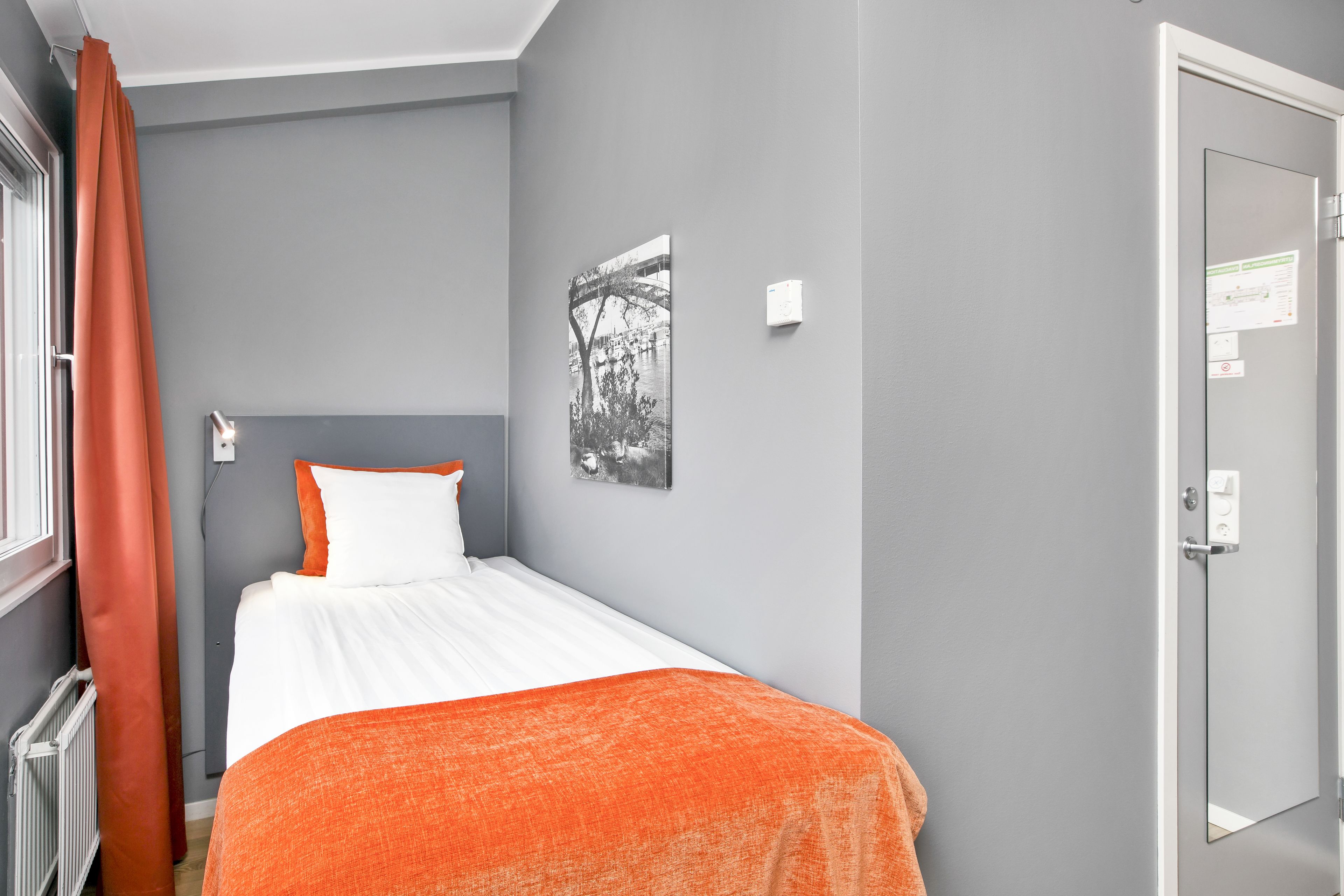 Un letto rifatto con biancheria da letto bianca e arancione, una parete grigia con una fotografia in bianco e nero e una porta con uno specchio e un interruttore della luce.
