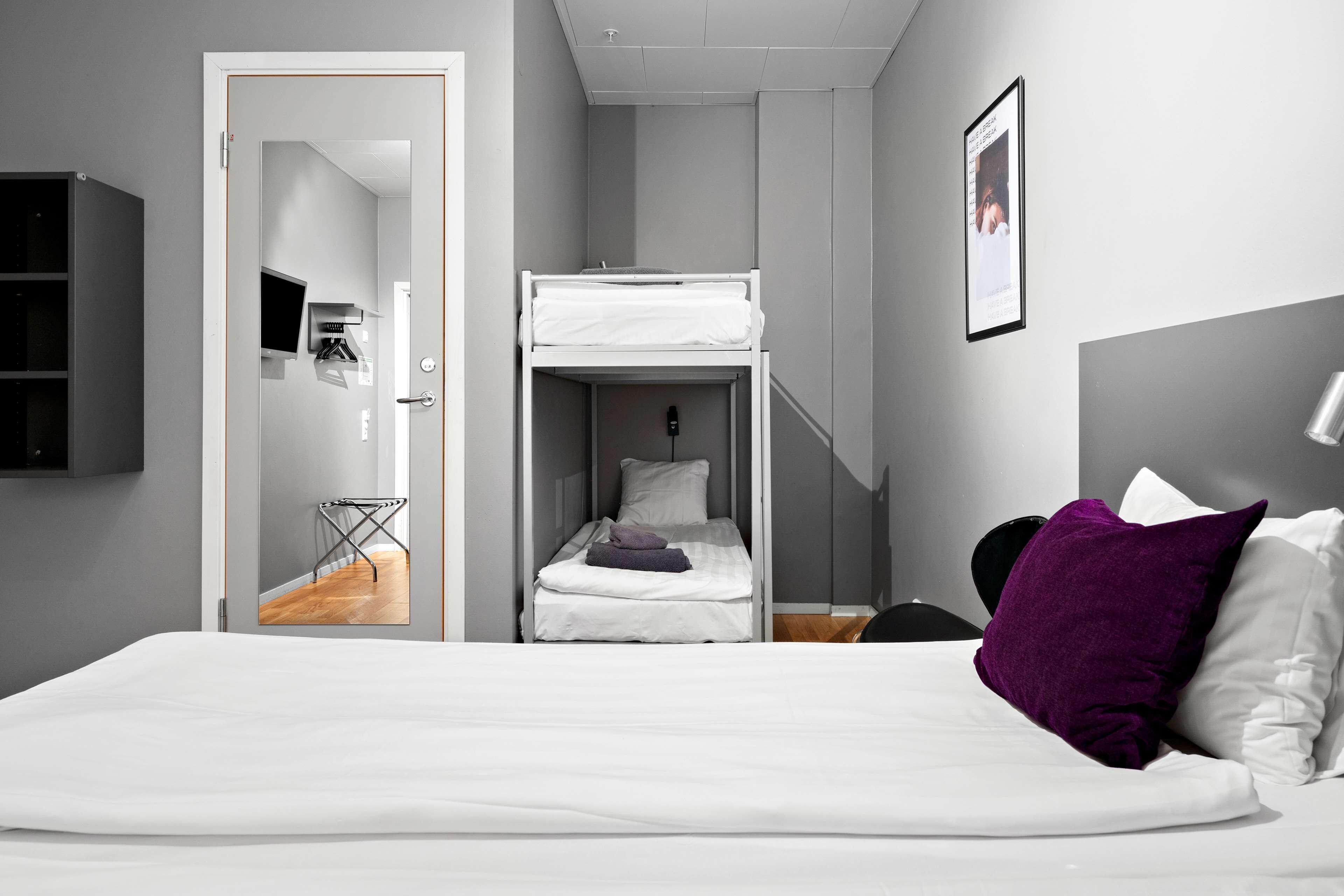 Habitación de hotel moderna con literas, televisor de pantalla plana, una mesita y cama con ropa de cama blanca y almohadas moradas.
