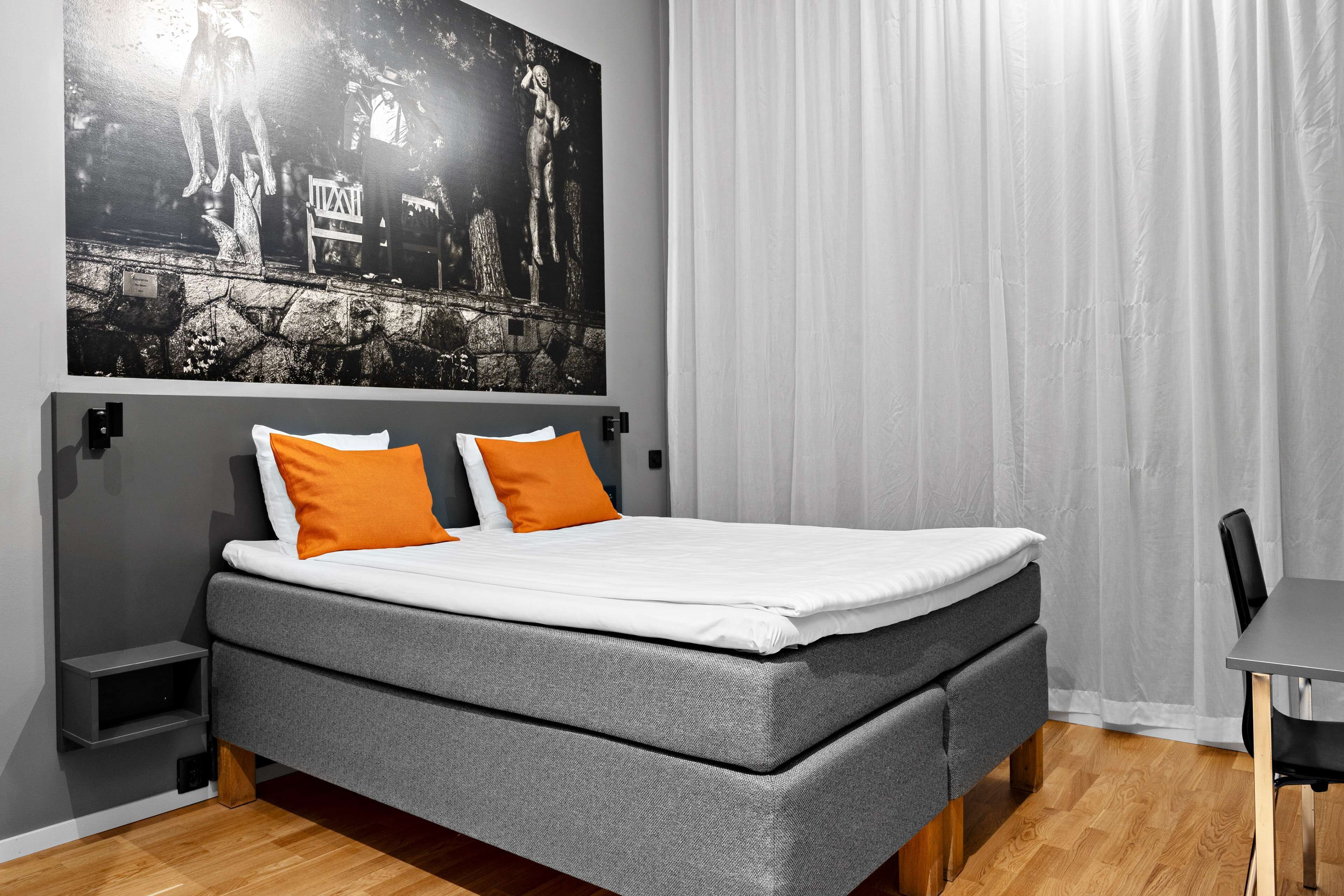 A modern bedroom with a grey sofa bed, two orange pillows, a white mattress, a black and white artwork on the wall, and a wooden floor.