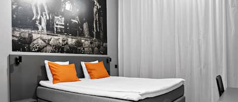 A modern bedroom with a grey sofa bed, two orange pillows, a white mattress, a black and white artwork on the wall, and a wooden floor.