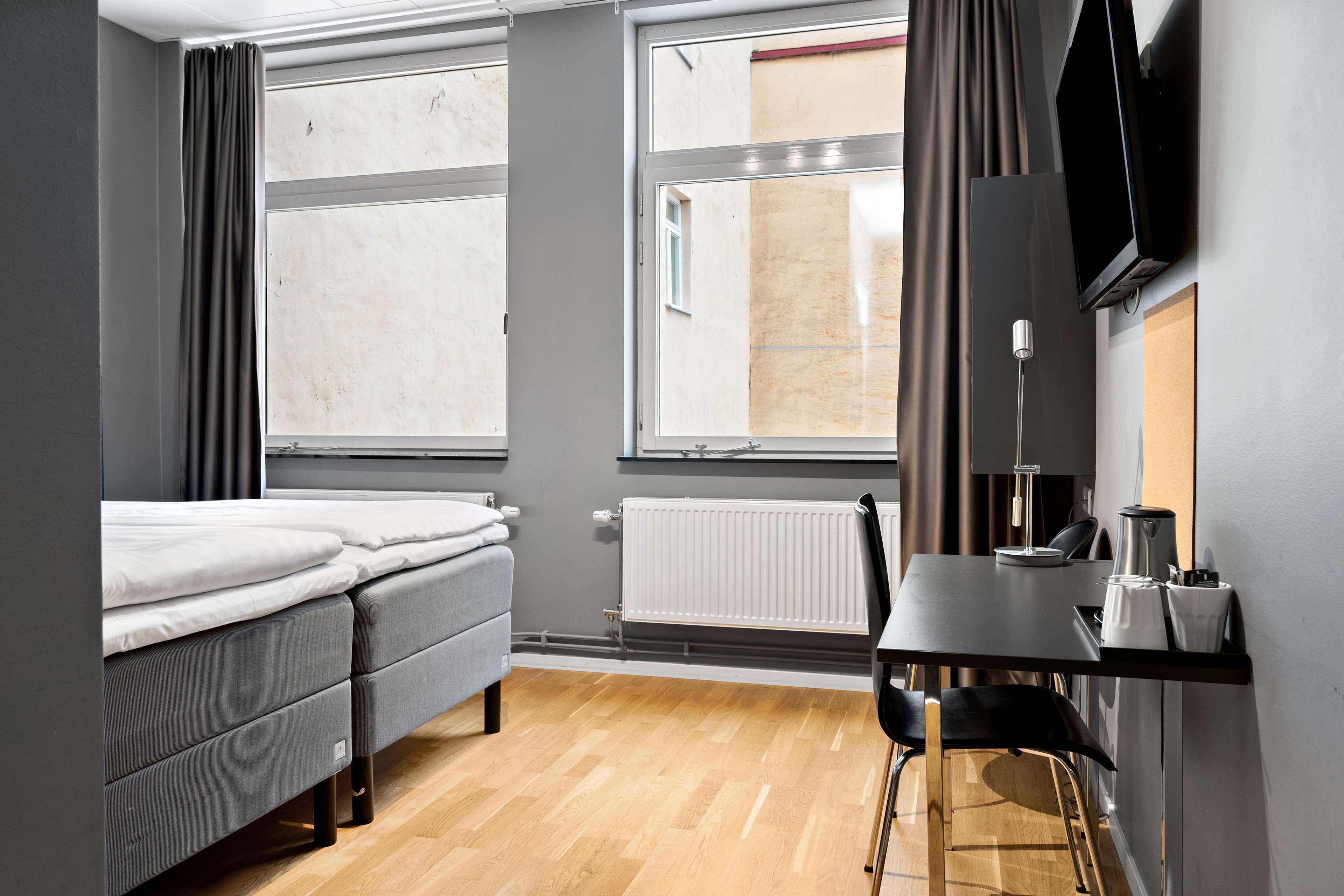 A compact hotel room with a bed, a desk, a chair, a TV, and large windows.