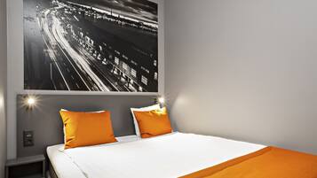Habitación de hotel moderna con una cama grande, dos almohadas naranjas, una colcha blanca y una vista nocturna de la ciudad en la pared.