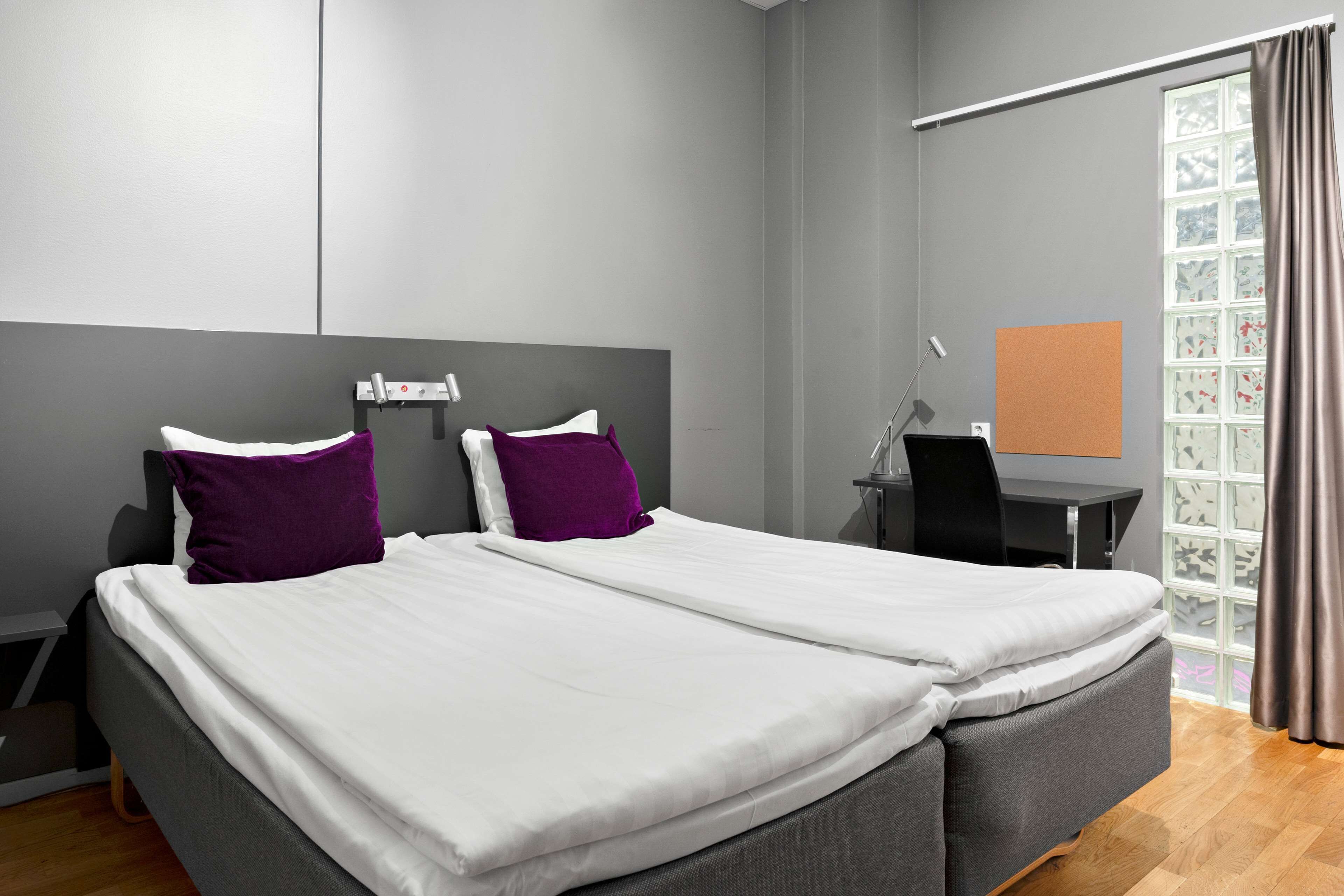 Una habitación de hotel moderna con una cama grande, dos almohadas moradas, un escritorio y una ventana con cortinas.