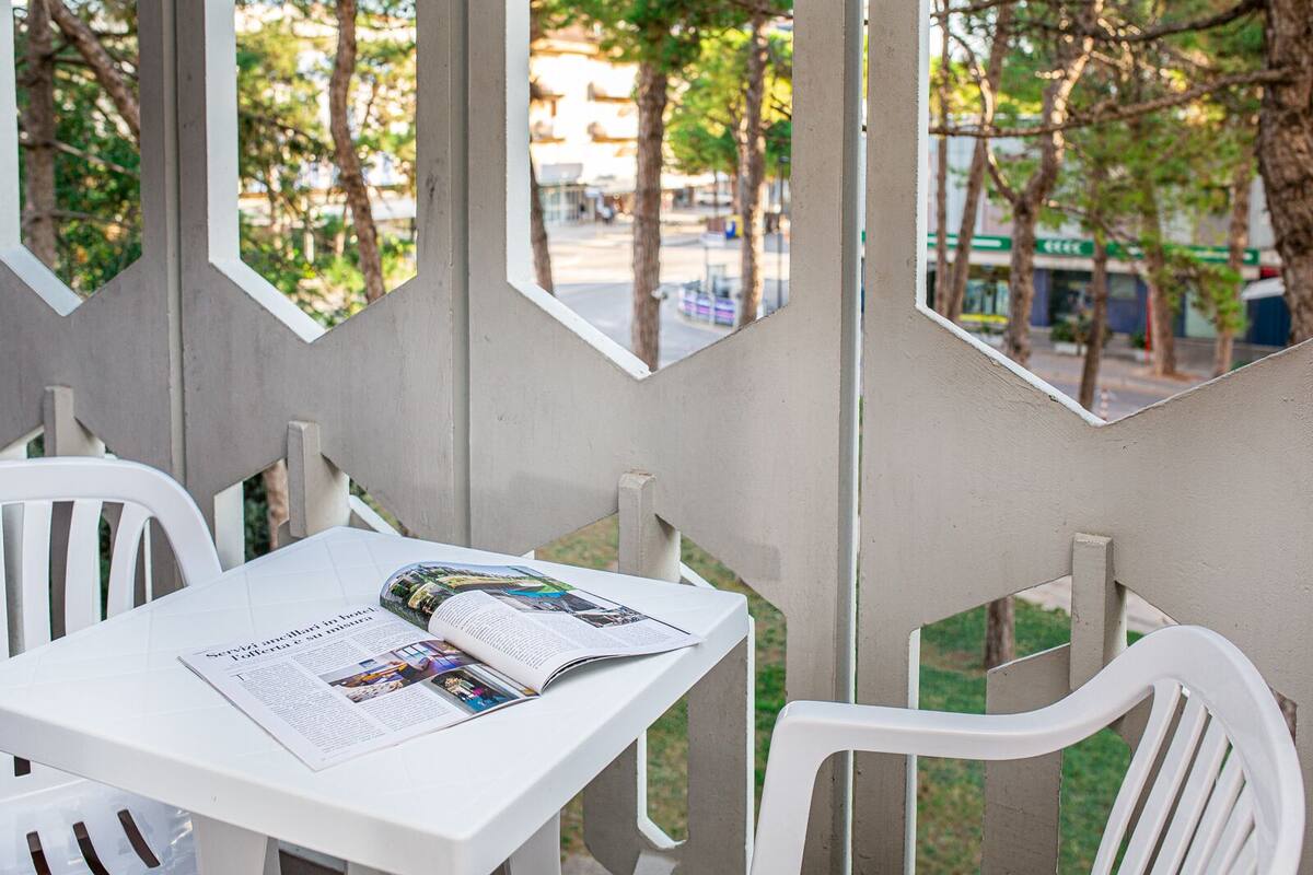 A white table with a magazine and a book, surrounded by white chairs, in a sheltered outdoor area with trees and a street view.