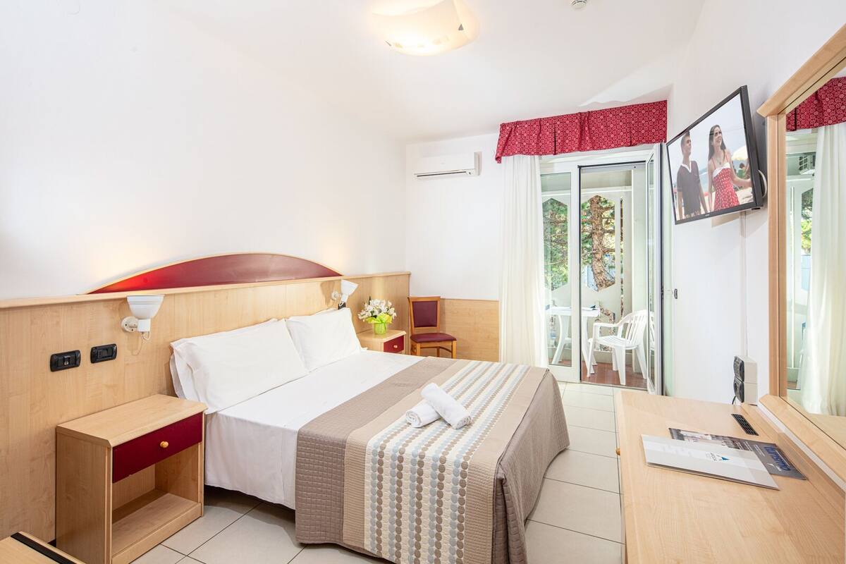 A hotel room with a bed, bedside table, a desk with a chair, a TV, and a sliding glass door leading to an outdoor area.