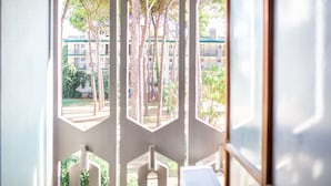 A view through a window with a geometric pattern, revealing a building with multiple floors and balconies, surrounded by trees and greenery.