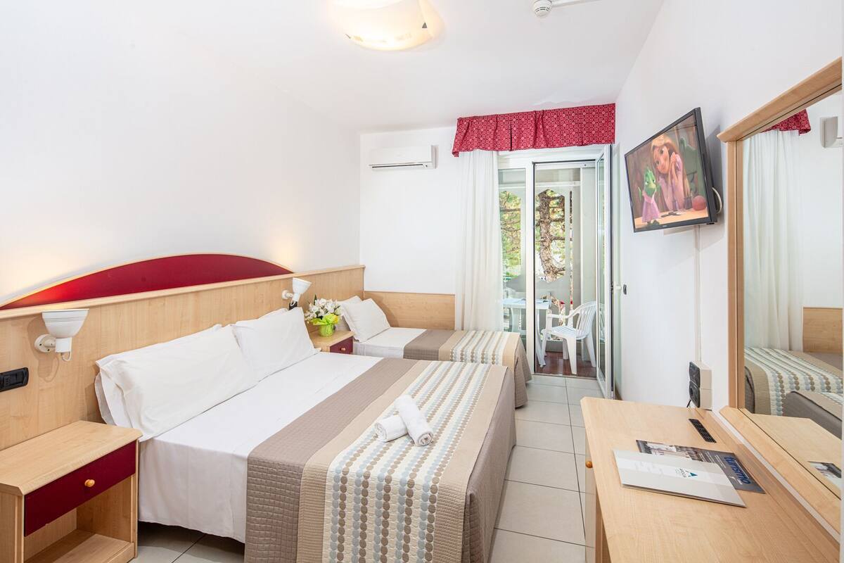 A hotel room with two beds, a desk, a chair, a TV, and a balcony with a table and chairs.