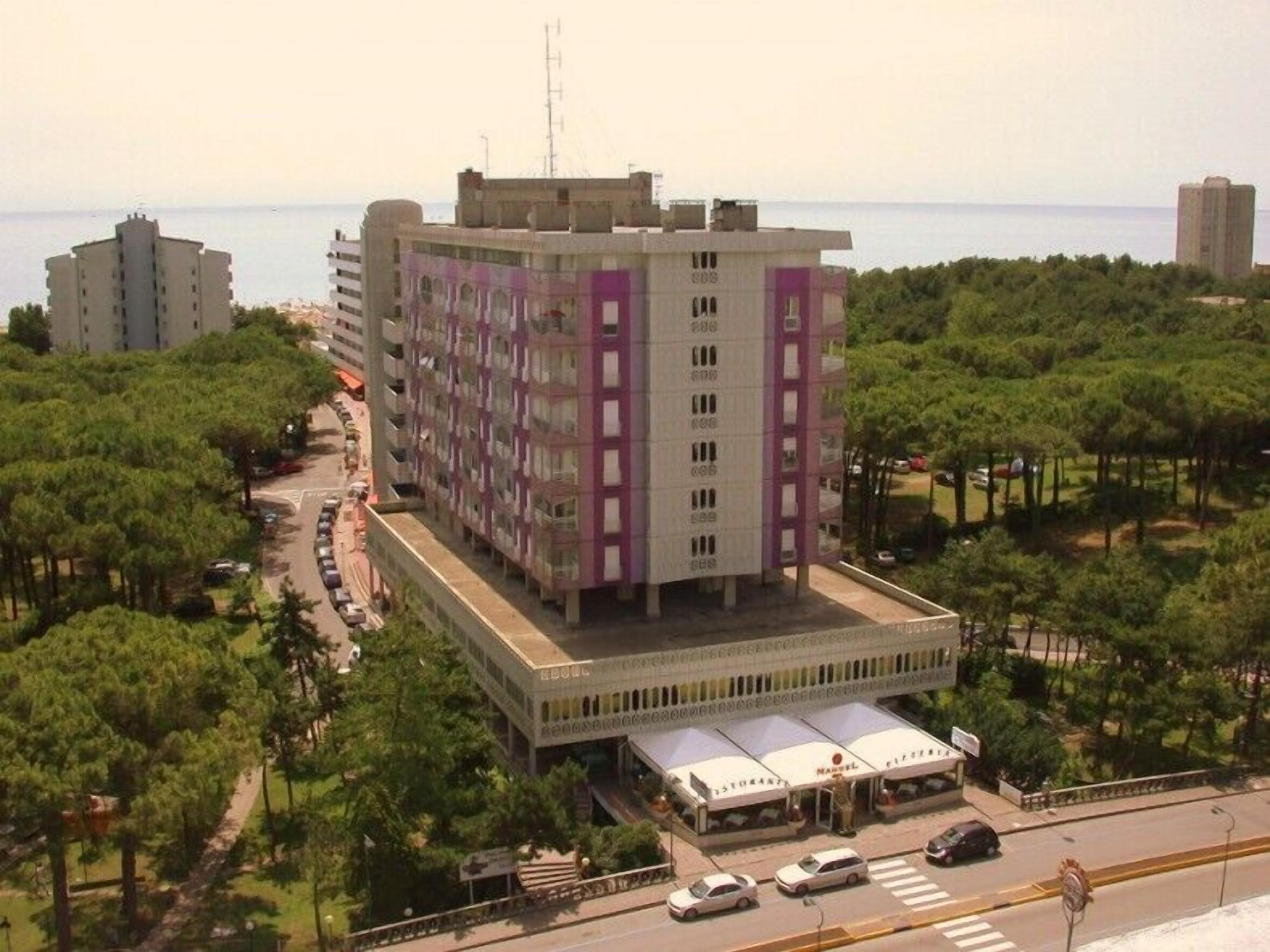 A multi-story hotel with a central courtyard, surrounded by trees and a view of the sea.