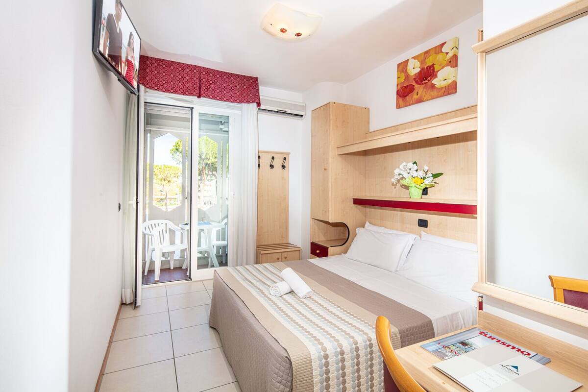 A hotel room with a bed, a desk with a chair, a wardrobe, and a sliding glass door leading to an outdoor area with chairs.