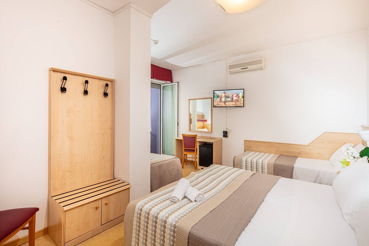 A hotel room with a wooden wardrobe, a desk with a chair, two beds with matching bedding, a TV on the wall, and a window with a view of the outside.