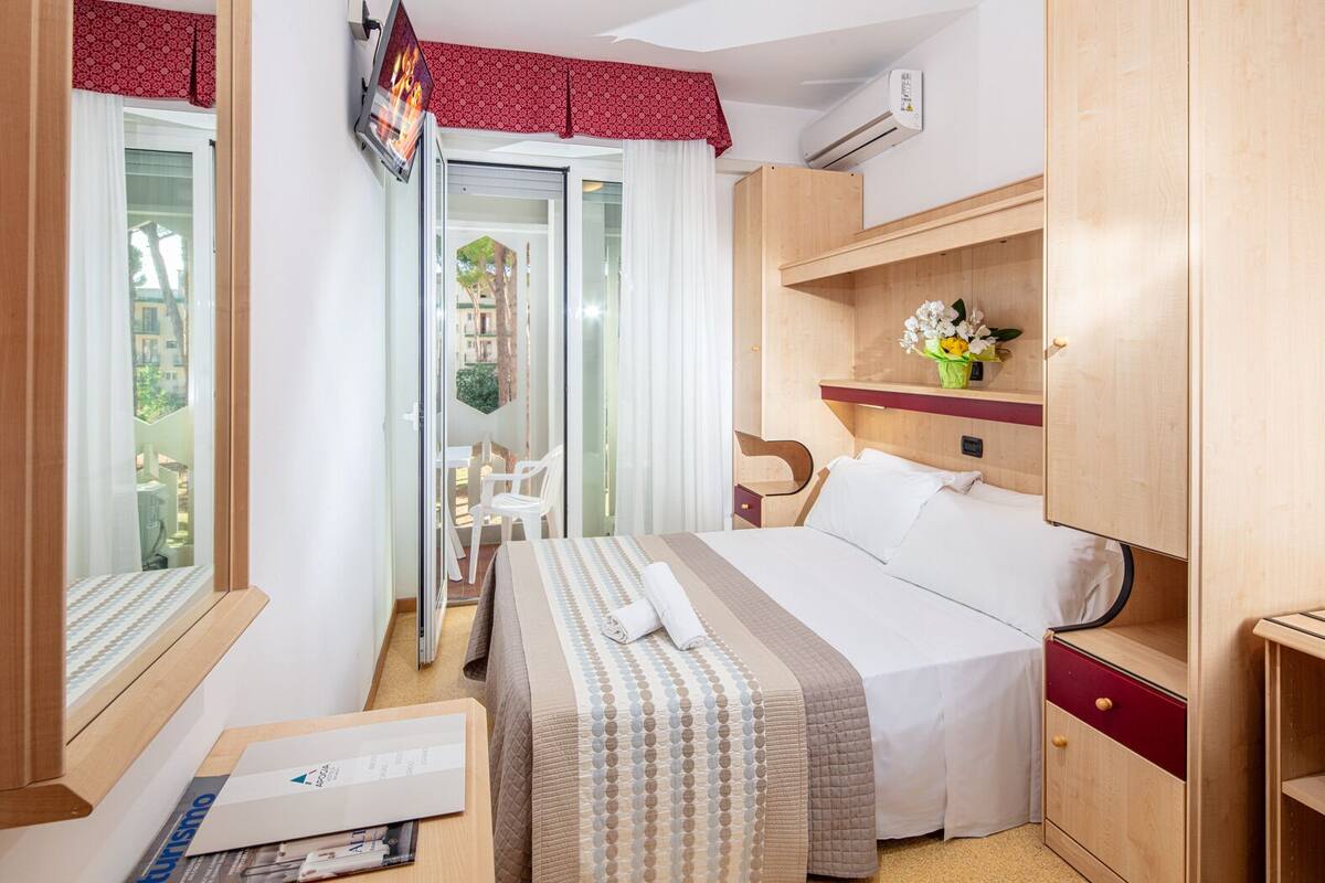 A hotel room with a bed, a desk, a chair, a TV, and a balcony with a view of buildings.