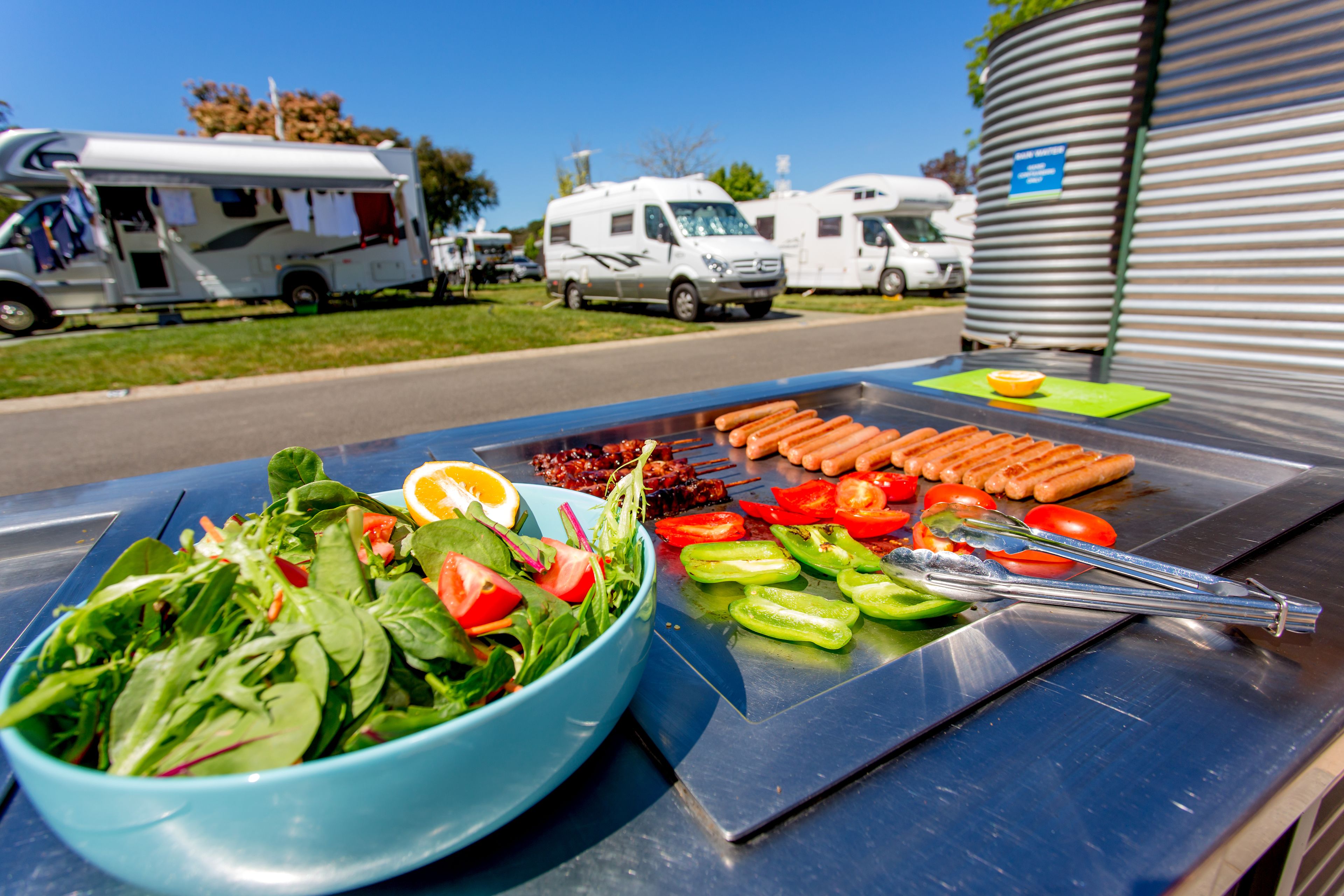Una parrilla con verduras y salchichas, un tazón de ensalada y vehículos recreativos al fondo.