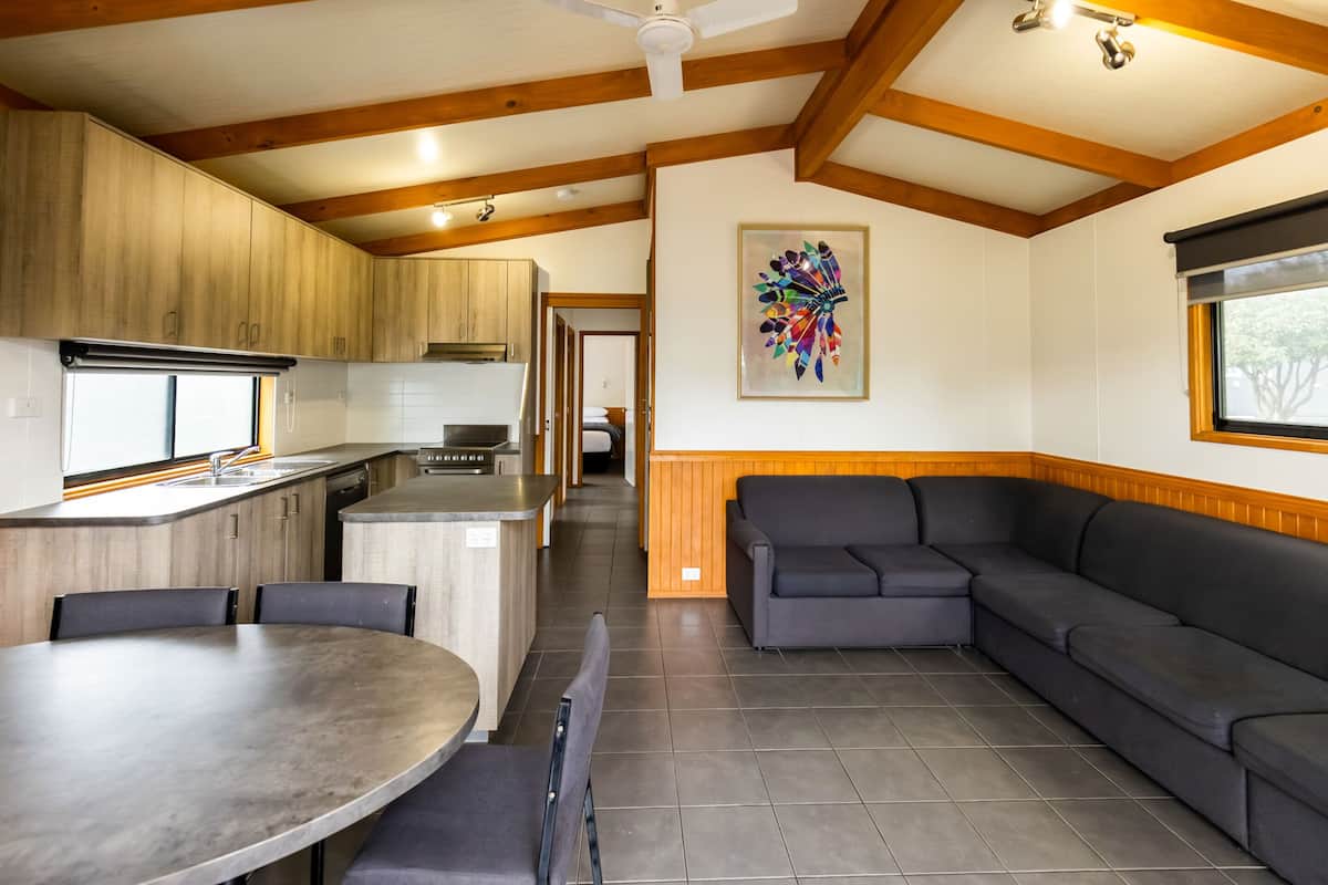 A modern kitchen with wooden cabinets, a dining area with a round table and chairs, a gray sofa, and a colorful abstract painting on the wall.