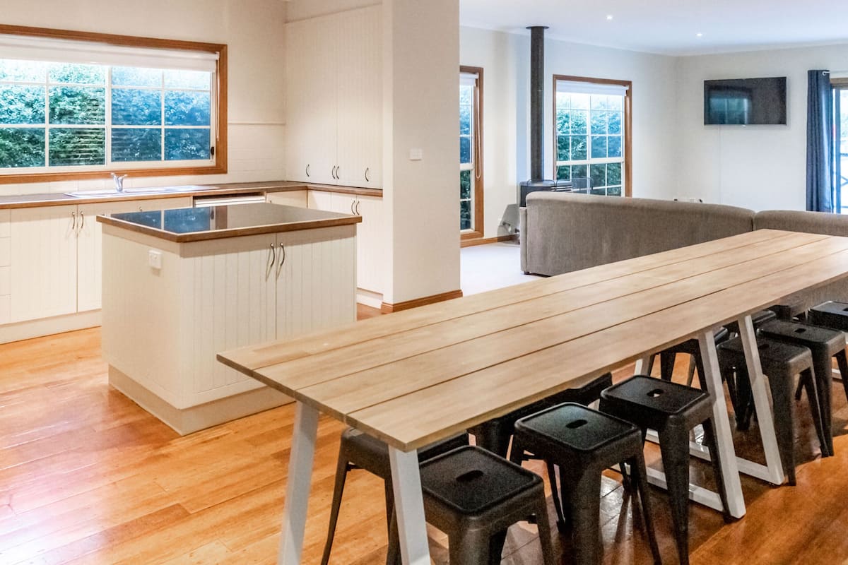 A modern kitchen with a wooden countertop and bar stools, a dining table, and a large window overlooking a forested area.