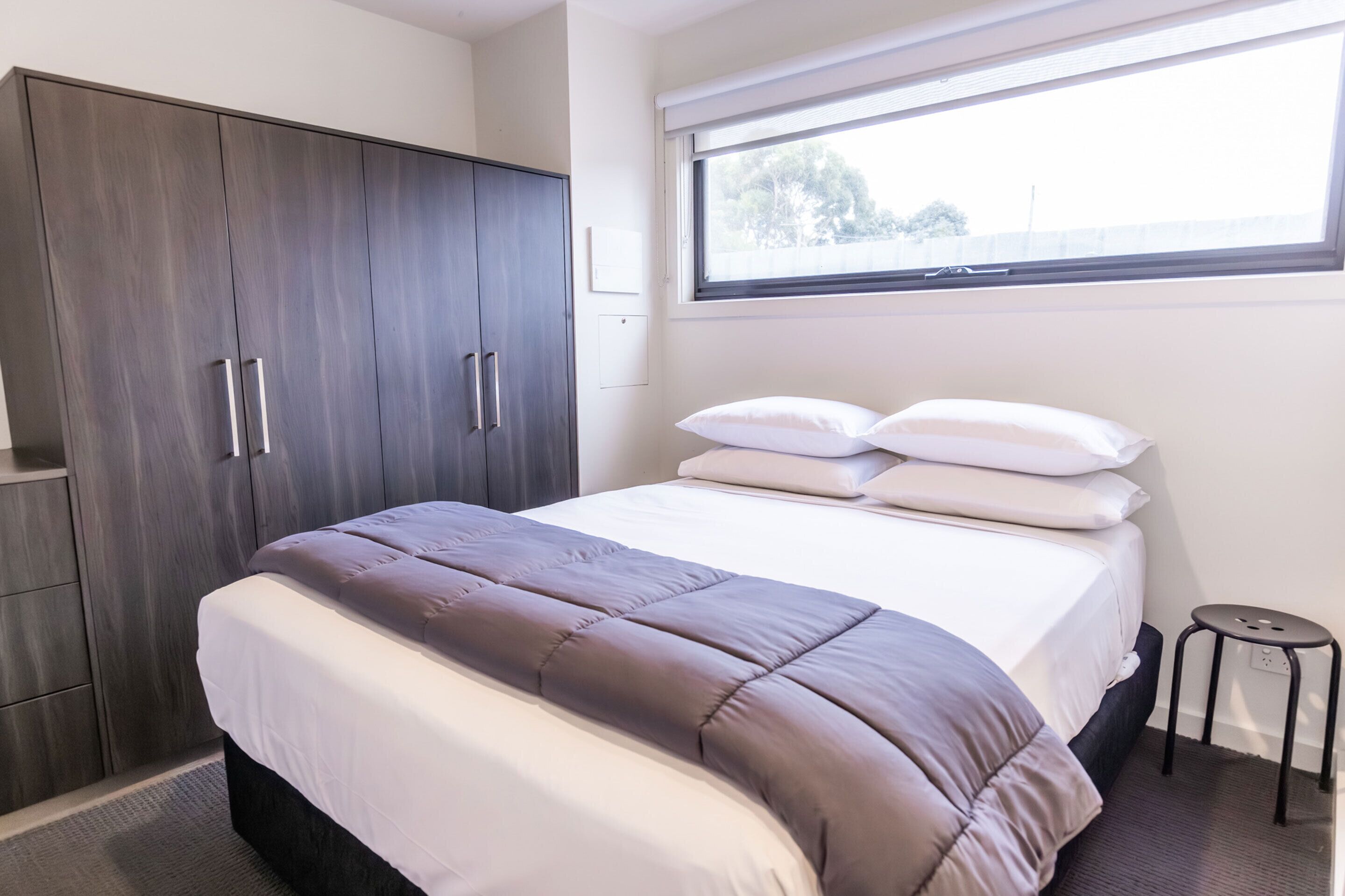 A bedroom with a bed, a wardrobe, a window, and a small stool.