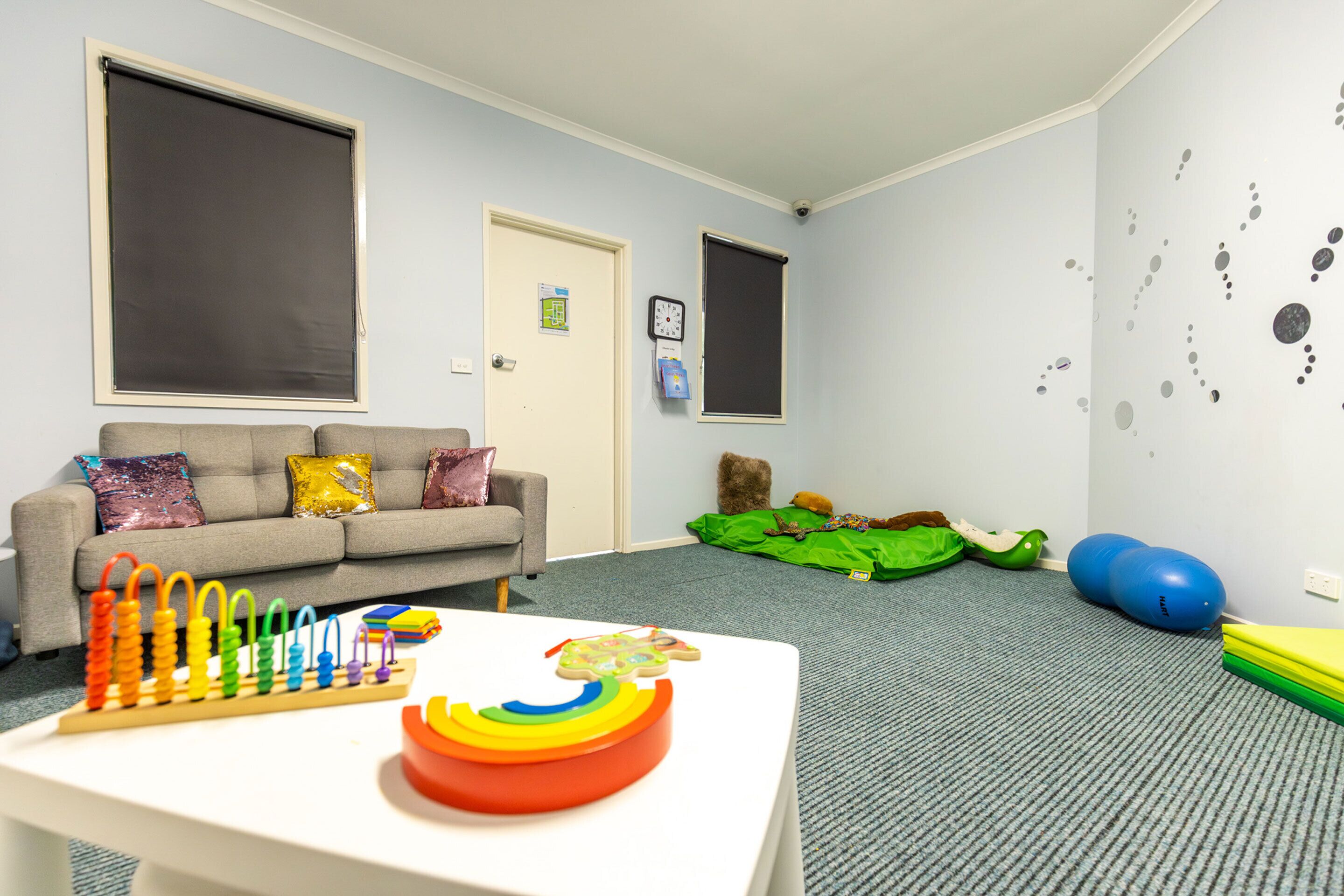 A room with a grey sofa, a white table with a colorful toy, a grey carpet, and a wall with a clock and decorative stickers.