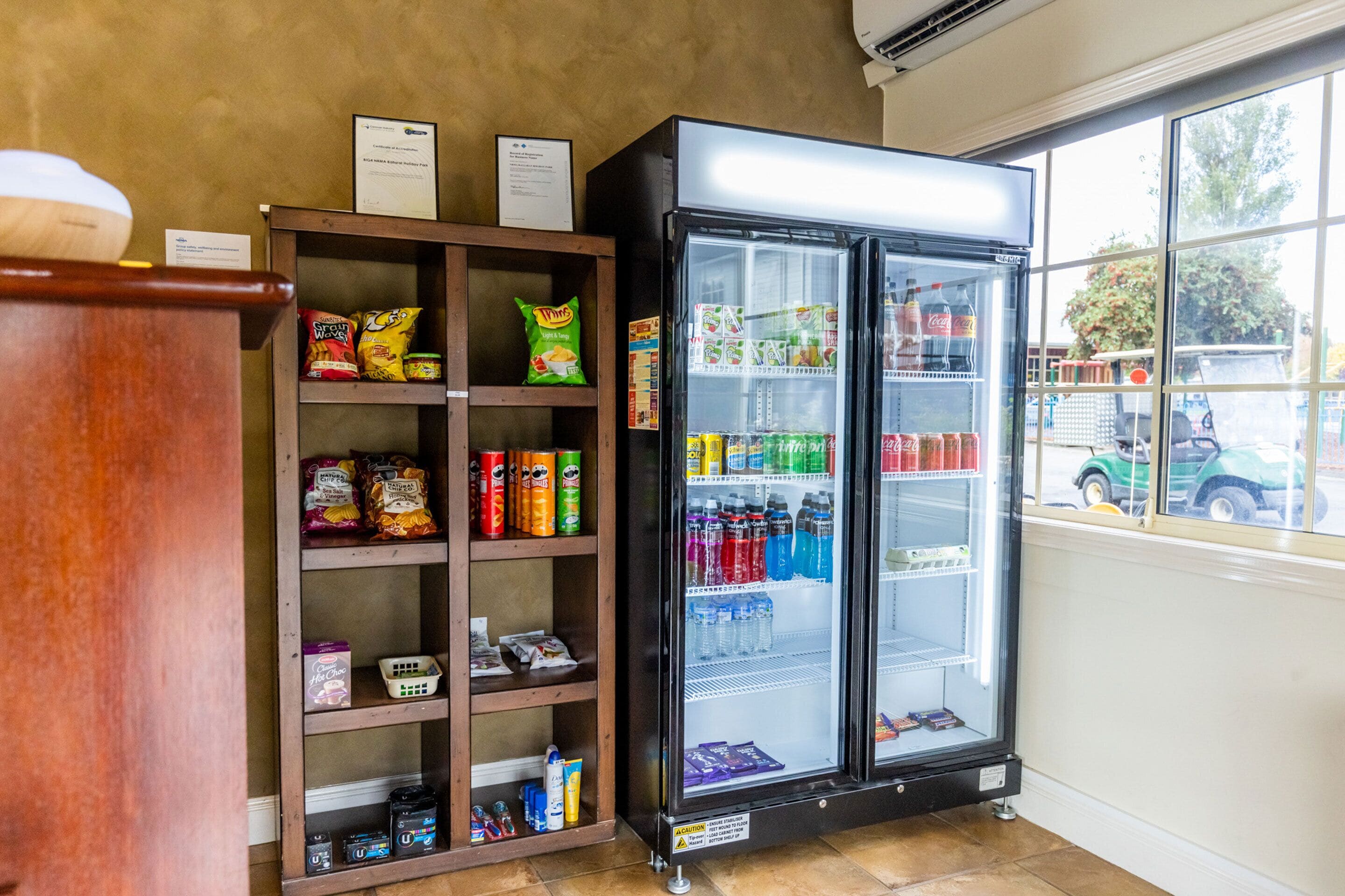 Máquina expendedora llena de bebidas y aperitivos, estante de madera con diversos alimentos, y ventana con vistas al exterior.
