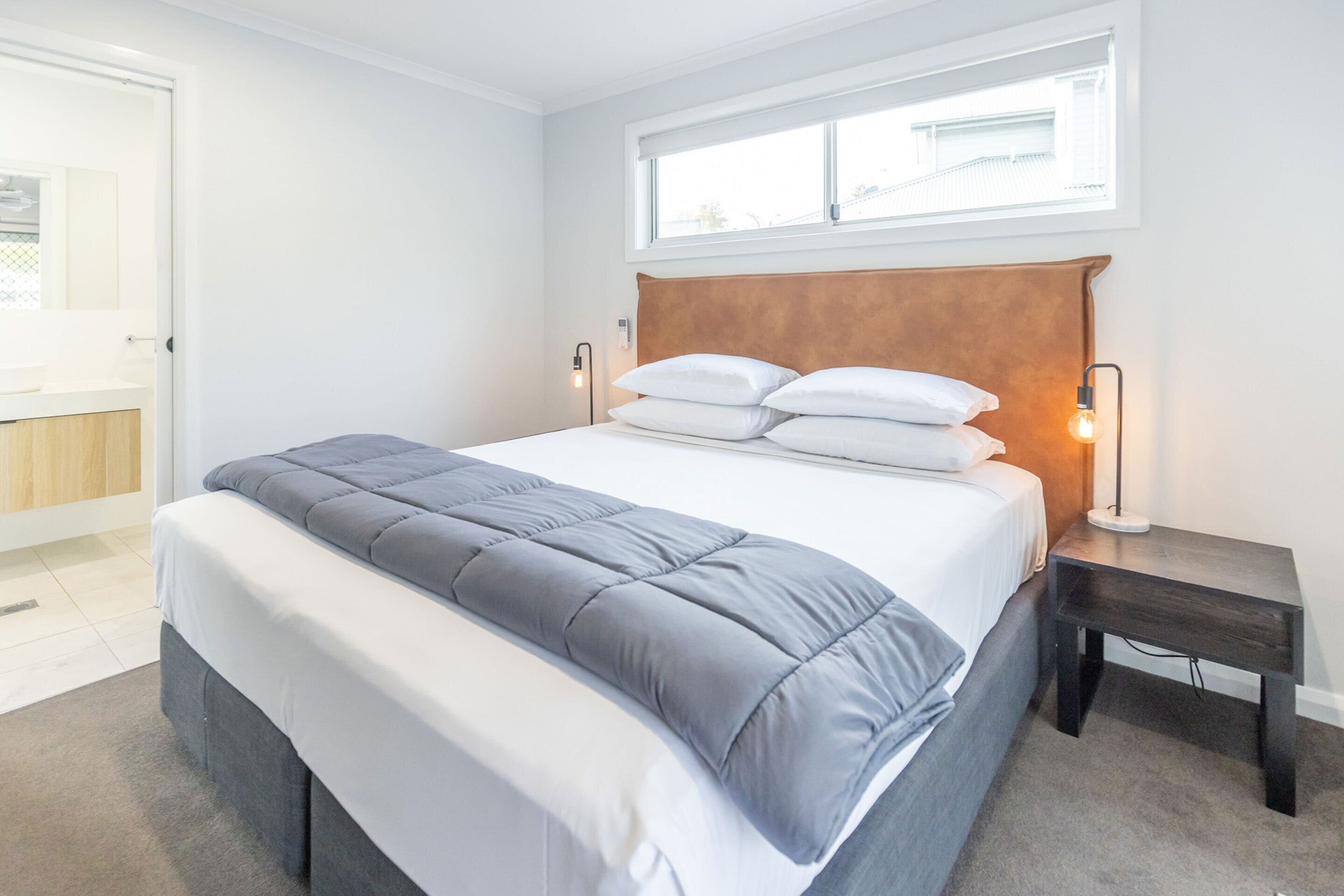 Une chambre moderne avec un grand lit, une tête de lit en bois, des lampes de chevet et une fenêtre avec des stores.