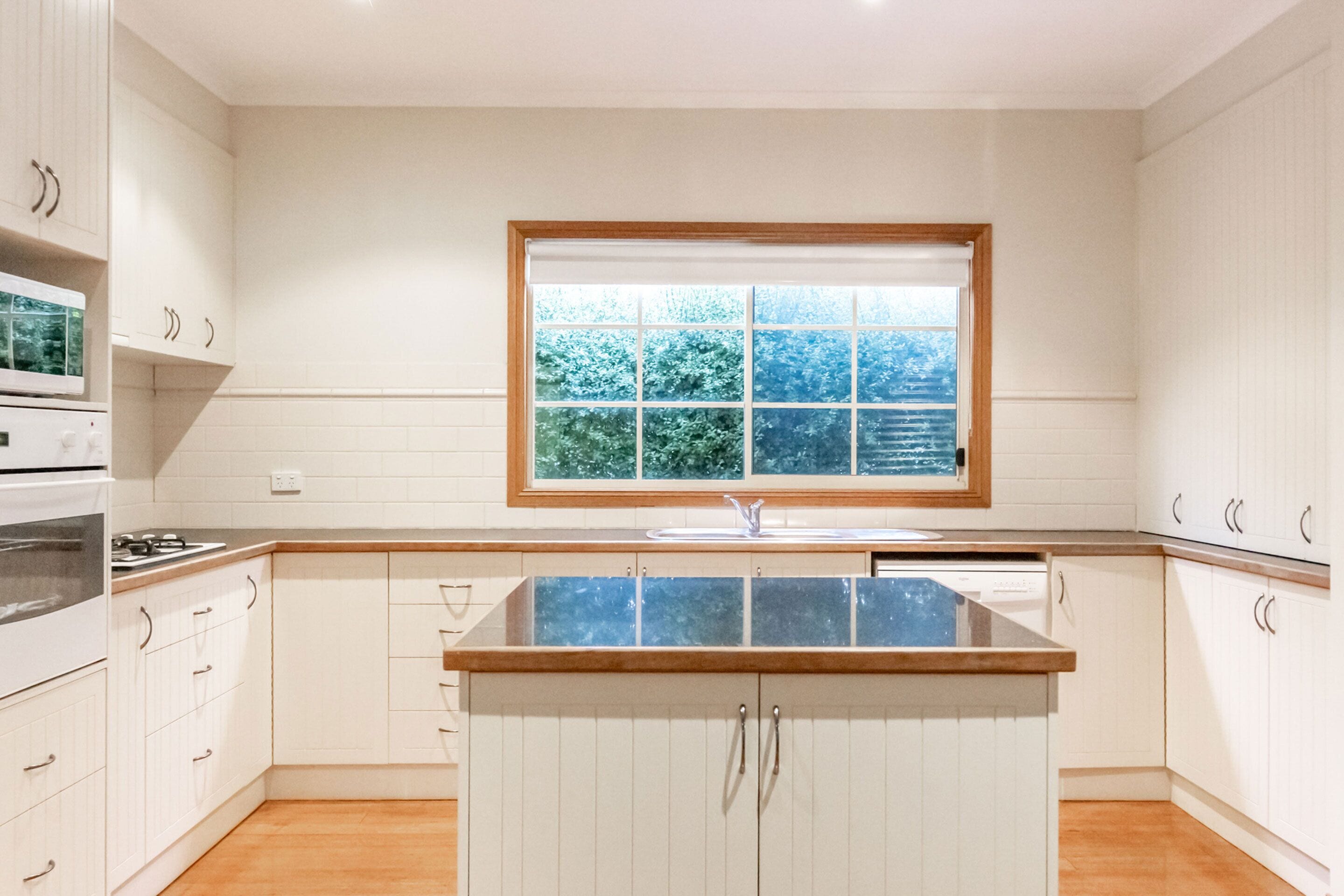 Una cocina moderna con armarios blancos, una isla central y un ventanal con vista a árboles.