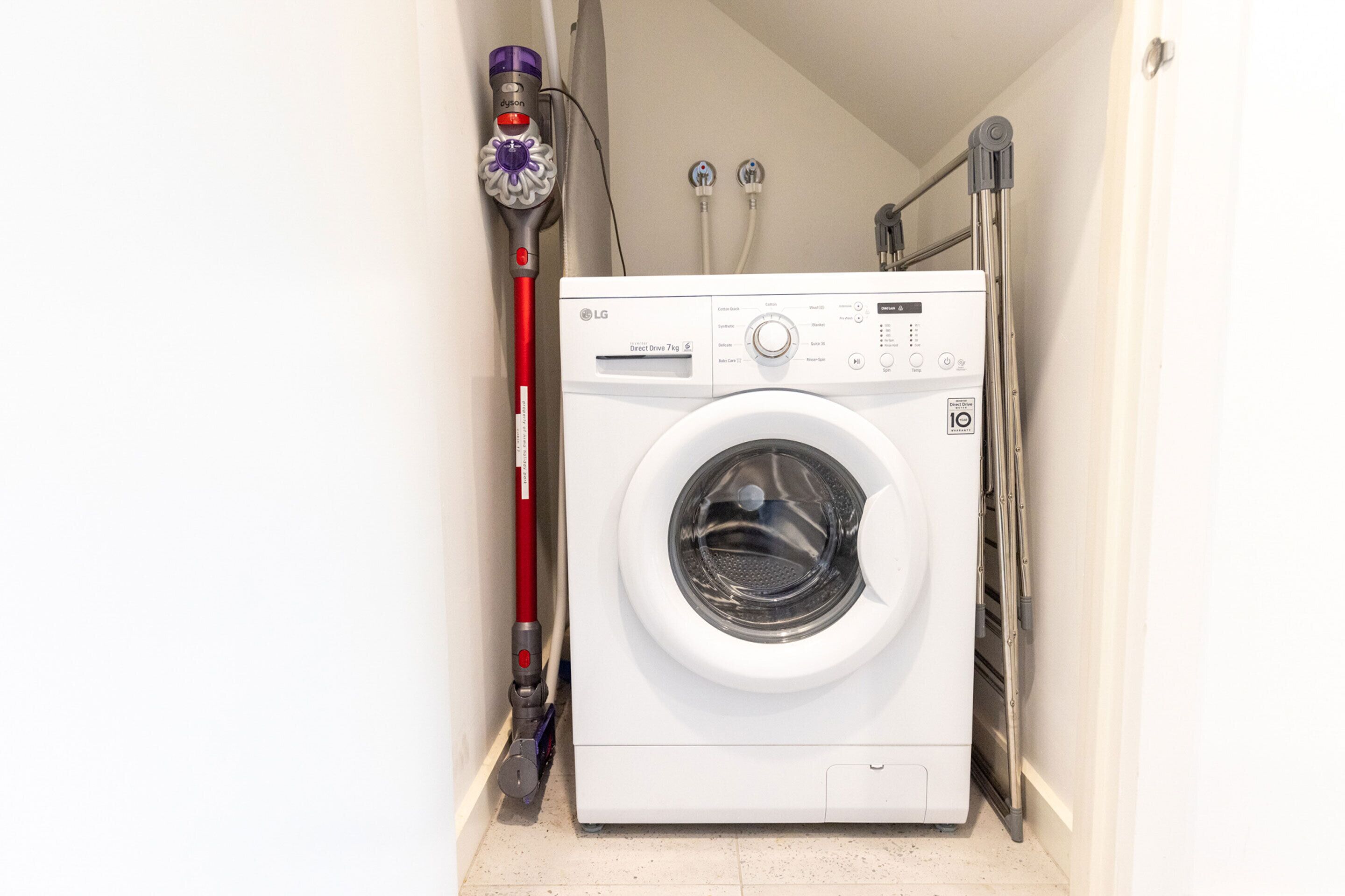 A compact washing machine in a narrow laundry room with a red mop and a vacuum cleaner on the wall.