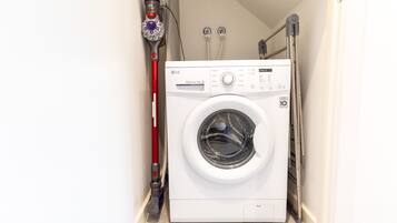 A compact washing machine in a narrow laundry room with a red mop and a vacuum cleaner on the wall.