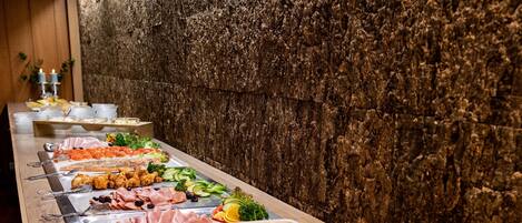 Buffet com diversas opções de comida, incluindo saladas, carnes e arroz, disposto sobre uma mesa comprida em frente a uma parede com textura.