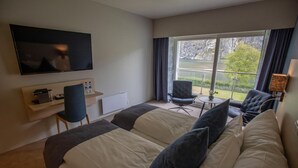 A hotel room with two beds, a TV, a small table with chairs, and a window with a view of a lake and trees. - Lærdal Hotel (Laerdal)